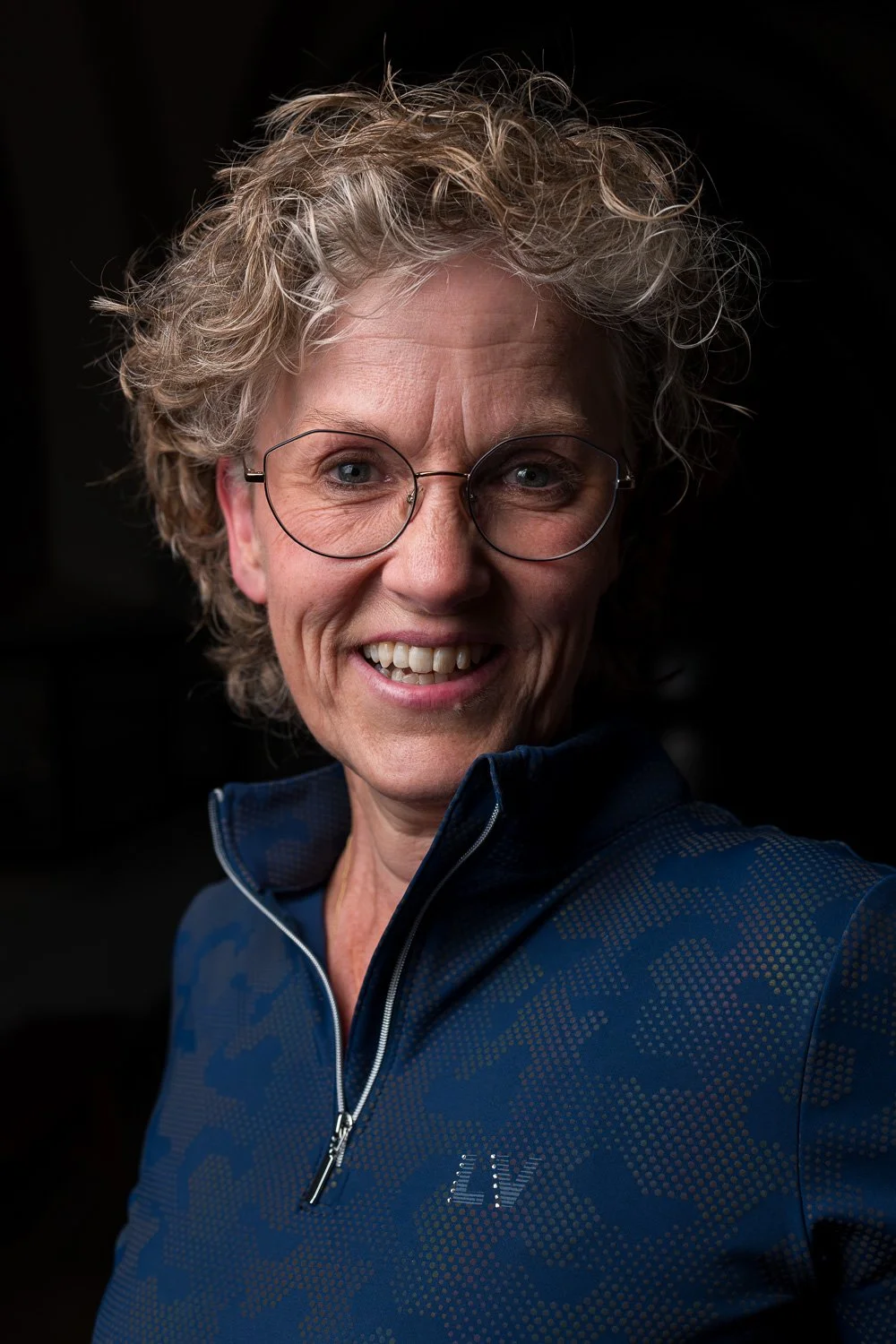 Portret van een lachende oudere vrouw met krullend grijs haar en ronde bril, gekleed in een blauwe sportjas met een wereldkaartpatroon. Donkere achtergrond.
