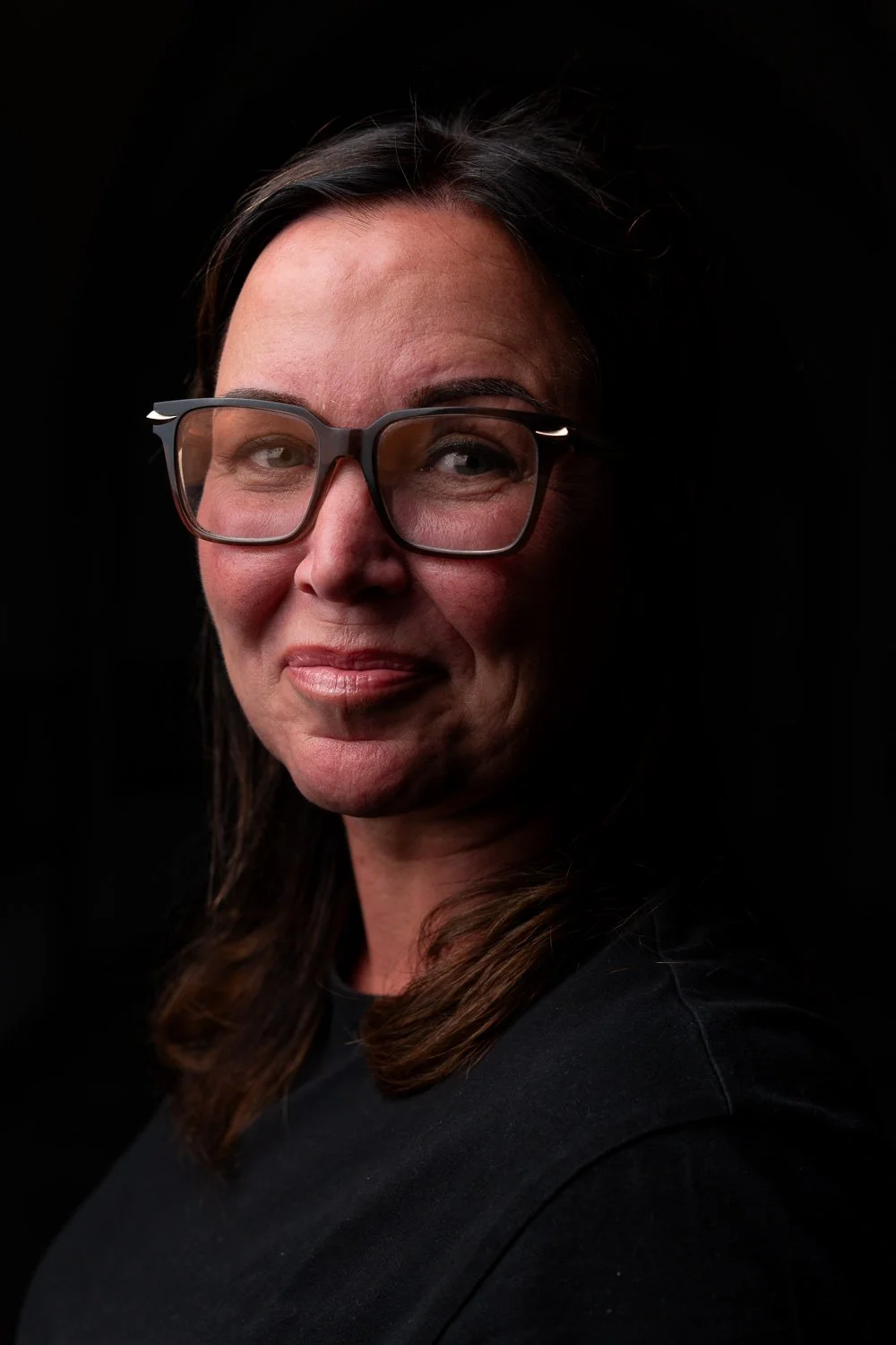 Portret van een vrouw met bruin haar, grote bril, lichte huid en een glimlach, tegen een zwarte achtergrond.