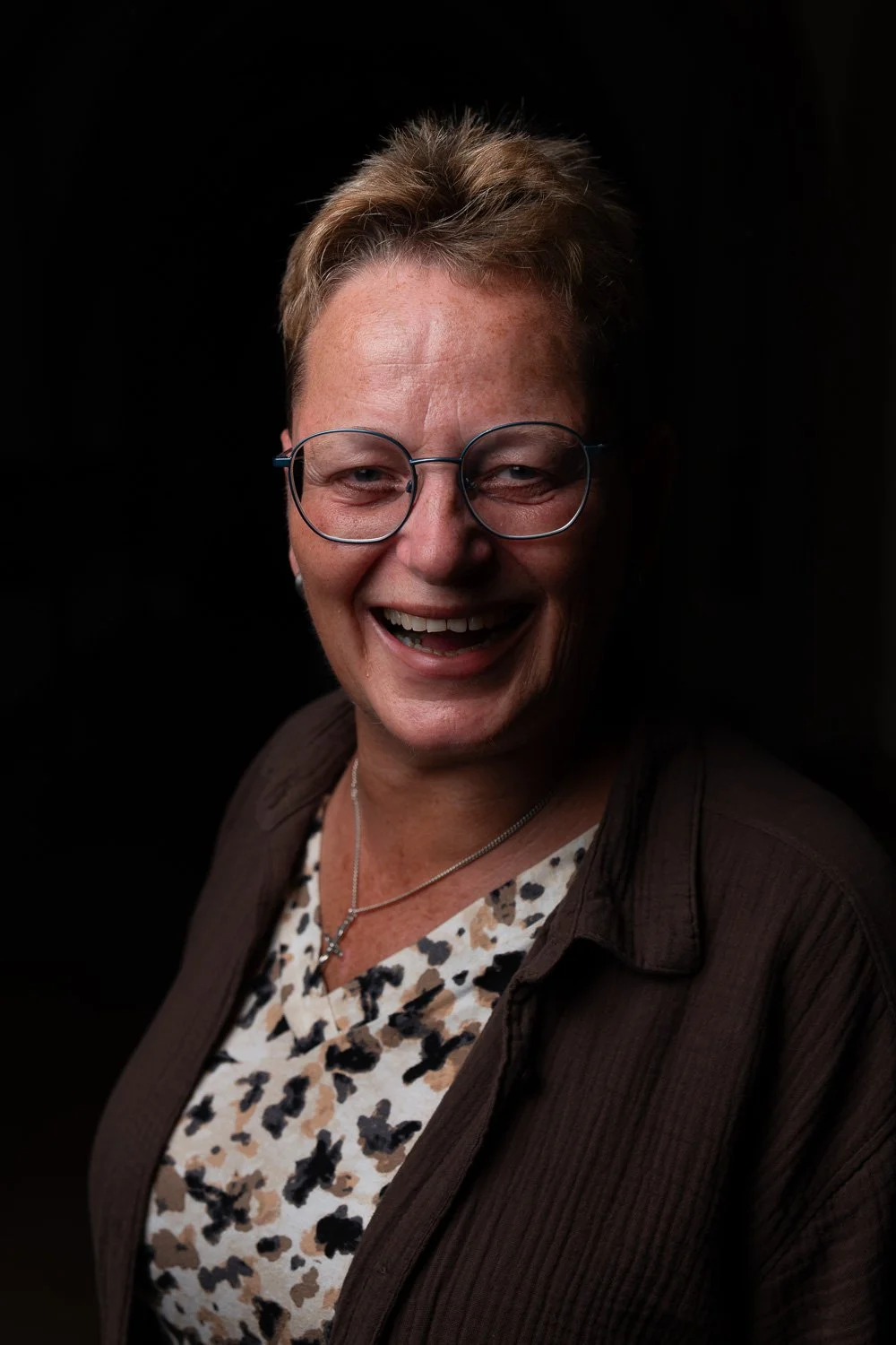 Zoom-in portret van een lachende vrouw met korte blond haar, bril en sieraden, tegen een zwarte achtergrond.