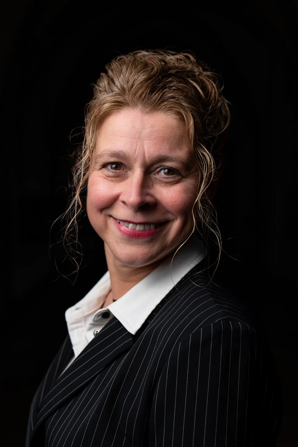 Portret van een lachende vrouw met lichtbruin, golvend haar, gekleed in een zwart met witte streepjes blazer en een witte blouse, tegen een zwarte achtergrond.