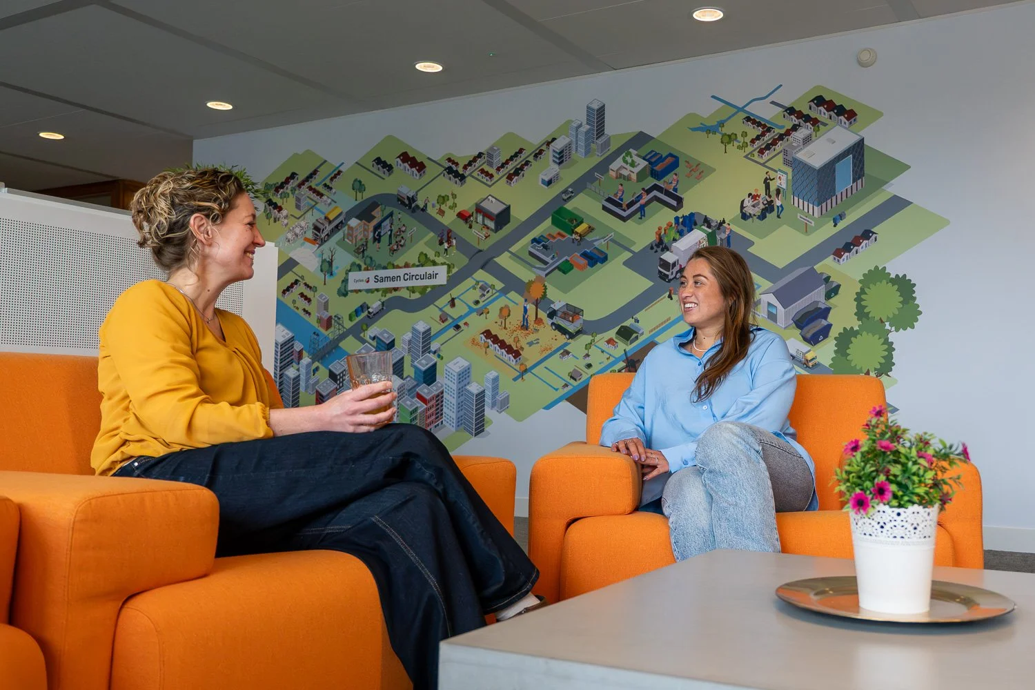Twee vrouwen die in een lobby of informele setting zitten, lachend en pratend, met een grote plattegrond of kaart van een stad op de achtergrond, en een tafel met bloemen voor zich.