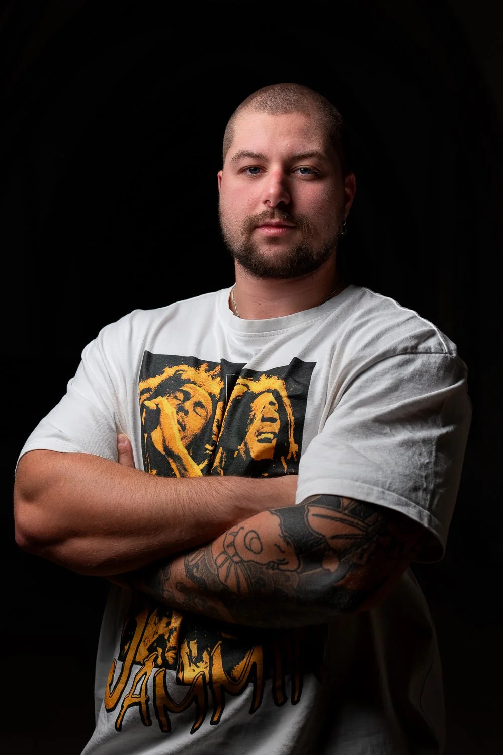 Portret van een man met een korte baard en een tattoo op zijn linkerarm, met armen gekruist, zittend voor een zwarte achtergrond, gedragen T-shirt met een gestileerde afbeelding van een vrouw.