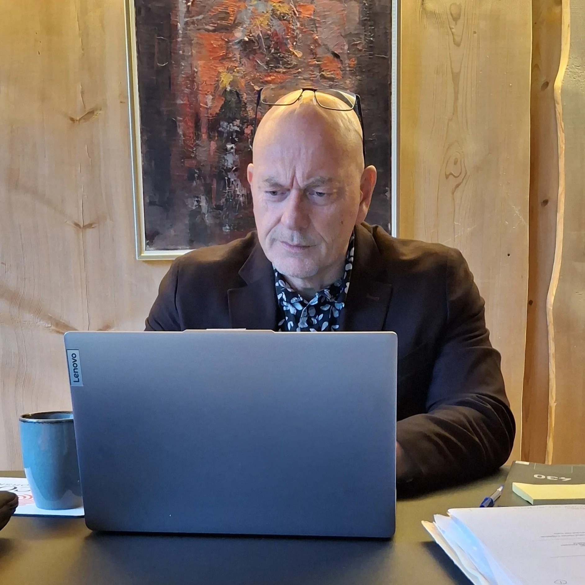 En mann med briller på hodet, som sitter foran en datamaskin, i et rom med trekk og et maleri på veggen bak ham.
