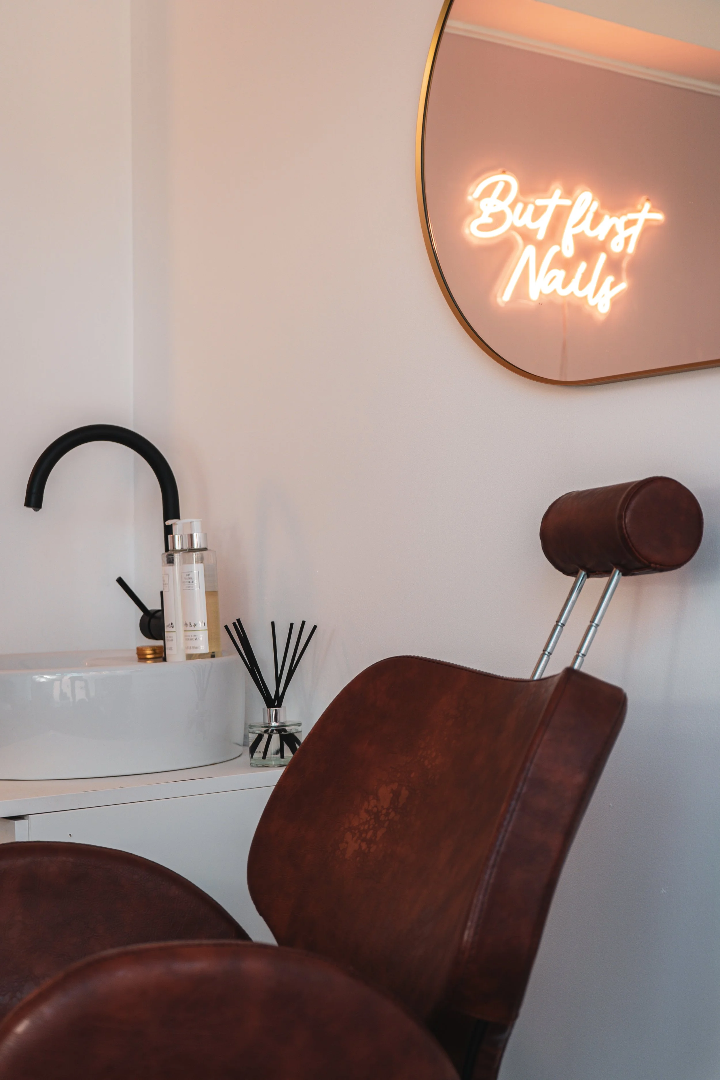 A bathroom decor scene with a white sink and black faucet, a bottle of liquid soap, a reed diffuser, a brown leather chair, and a wall mirror with neon sign reading 'But first, Nails'.