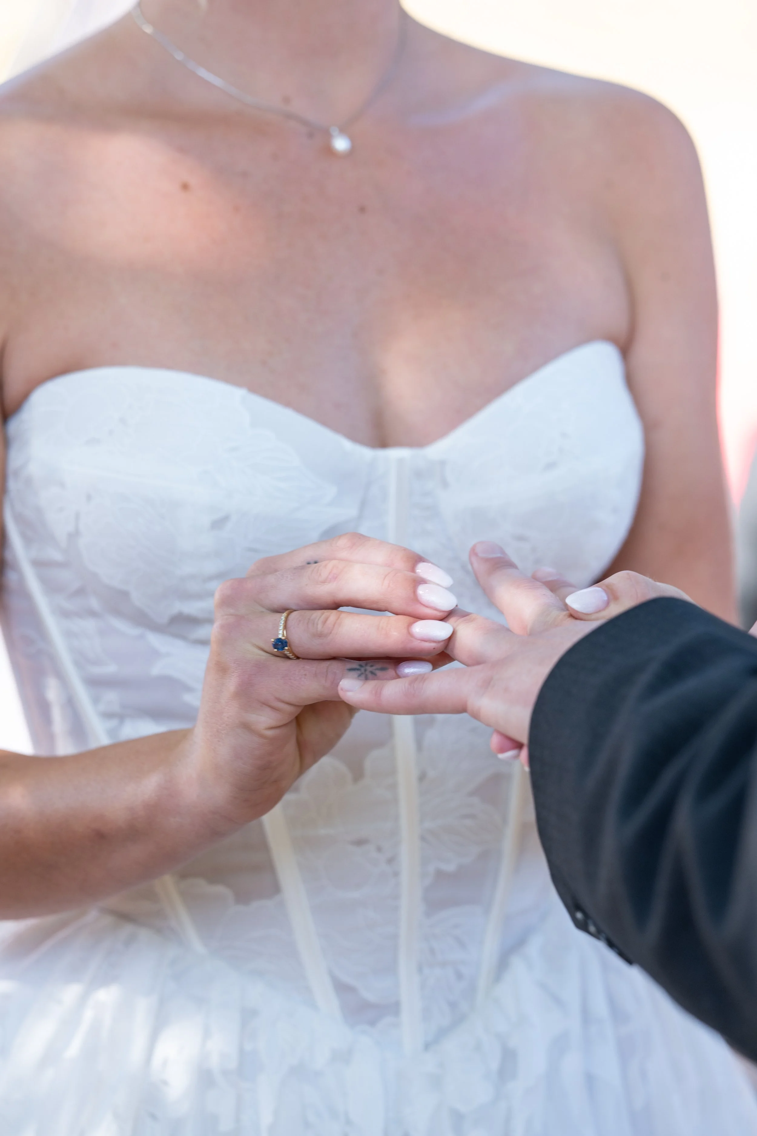 Close-up of bride and groom holding hands during wedding ceremony, bride wearing a white strapless dress with lace details and a delicate necklace, groom in a dark suit, outdoor setting with bright lighting.