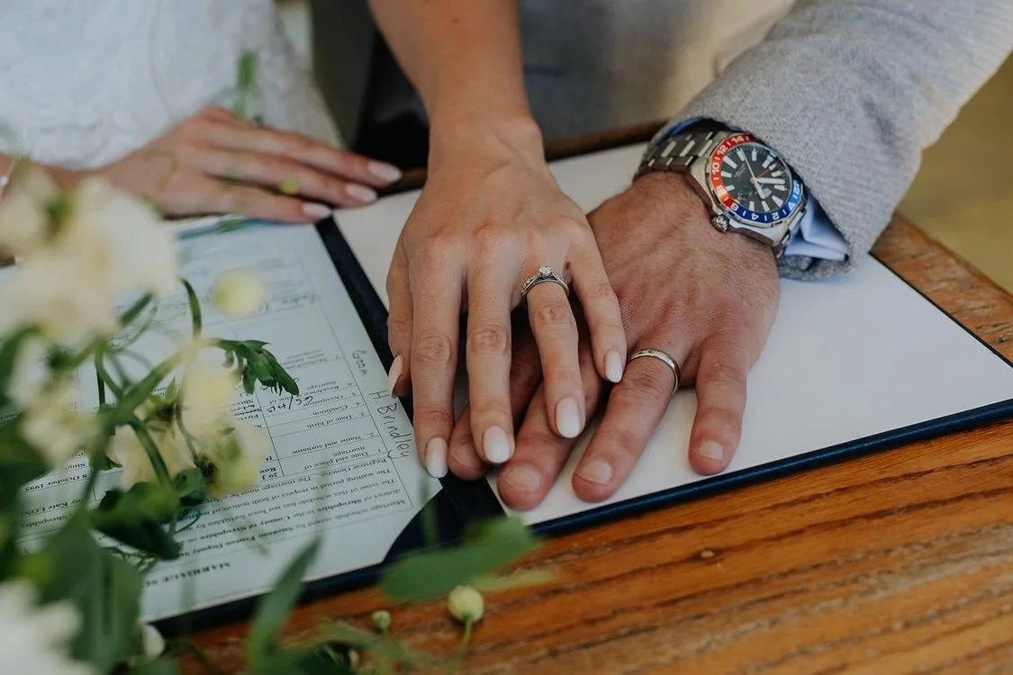 Close-up of a couple's hands with wedding rings, resting on a document on a wooden table, with flowers nearby.