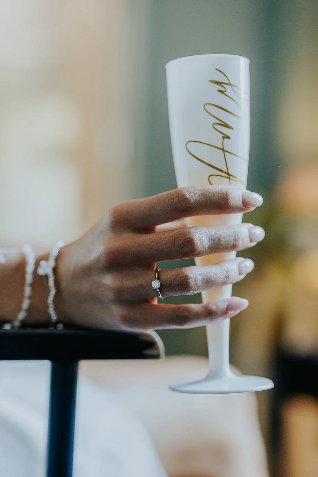 A hand with a diamond engagement ring and a pearl bracelet holding a white flute with gold script.