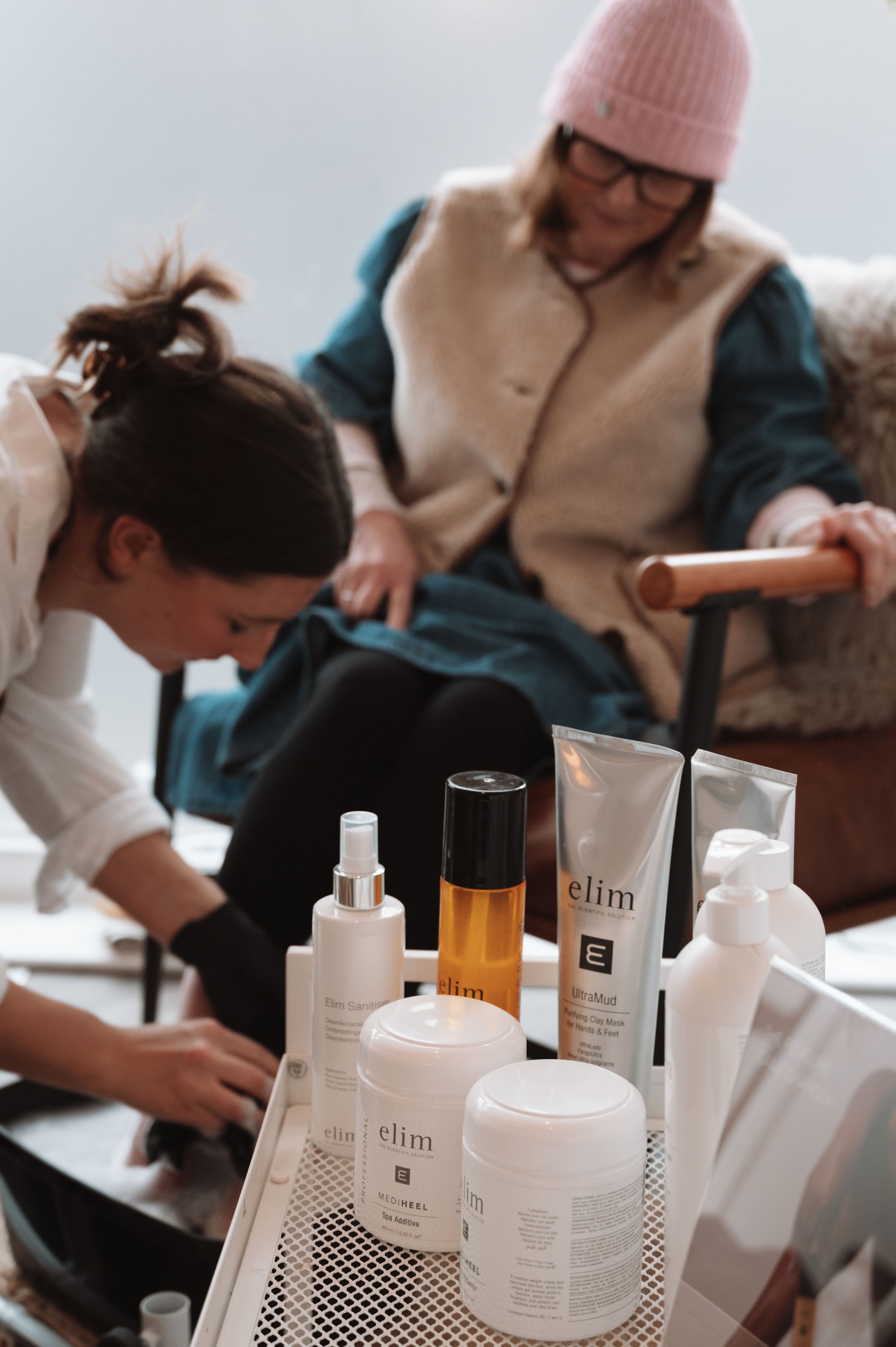 People working with skincare products and bottles on a white tray, with one person wearing glasses and a pink beanie in the background.
