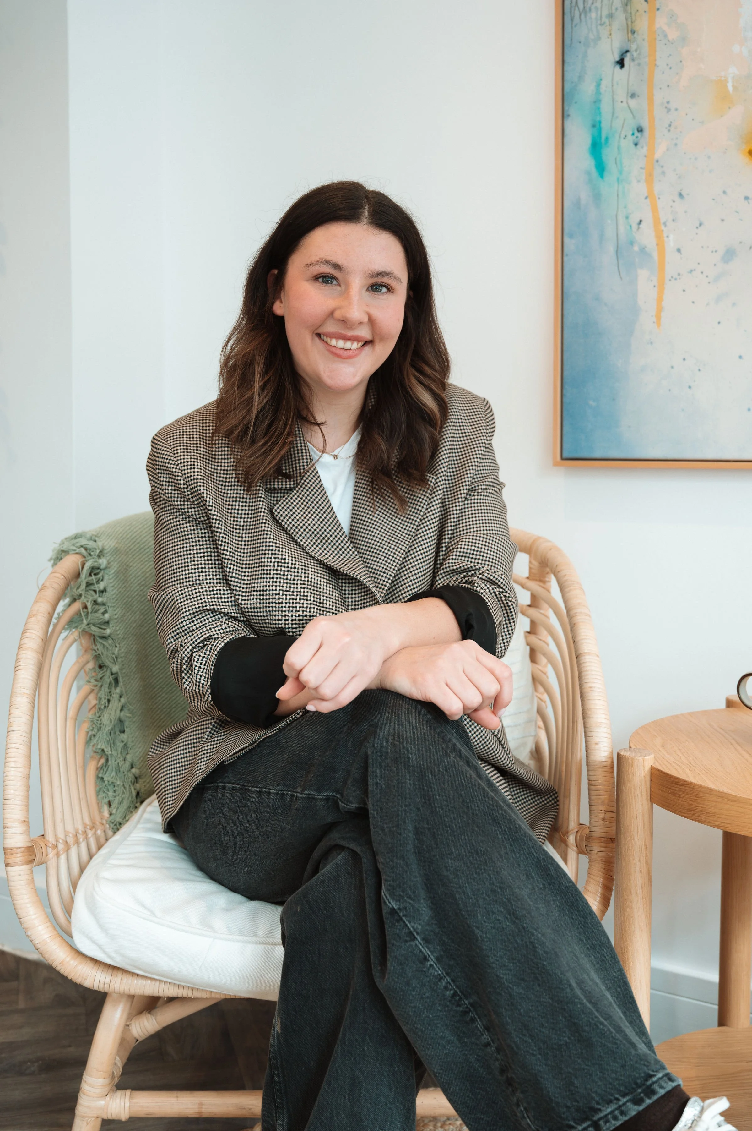 A woman with dark, shoulder-length hair and blue eyes is sitting on a white cushion on a light wood chair with a woven rattan frame. She is smiling and wearing a plaid blazer, black pants, and a white shirt. Behind her is a white wall with an abstract painting featuring blue, yellow, and beige colors. A round wooden side table is visible to her right.