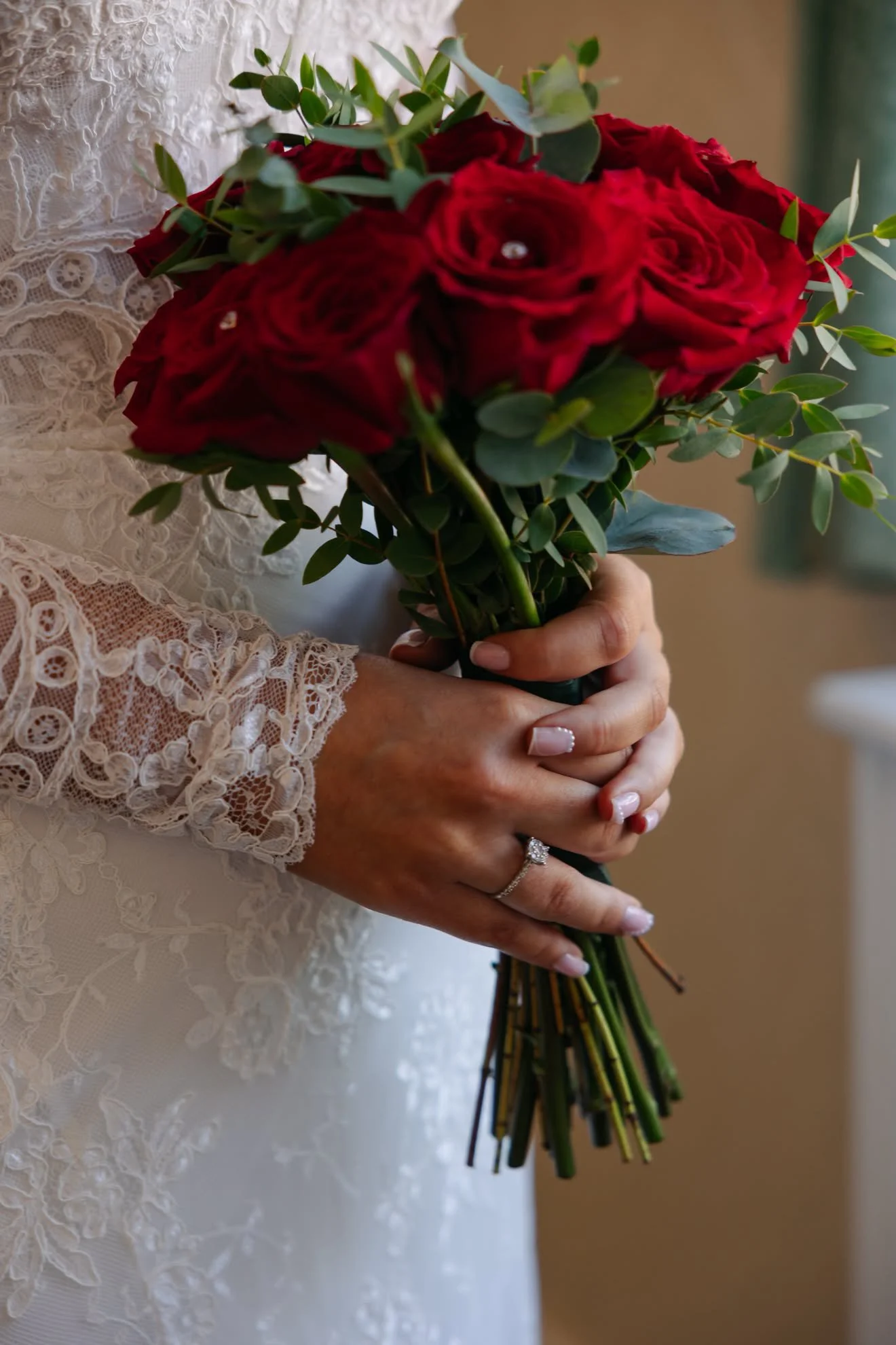 Close-up of a bride holding a bouquet of red roses and green leaves, wearing a white lace wedding dress with detailed embroidery.