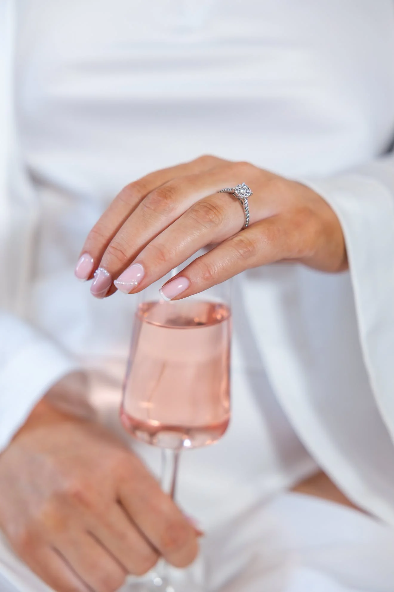 A person wearing a diamond engagement ring on their finger, holding a glass of pink-colored champagne or wine, with a blurred background of a white shirt and a hand in the foreground.