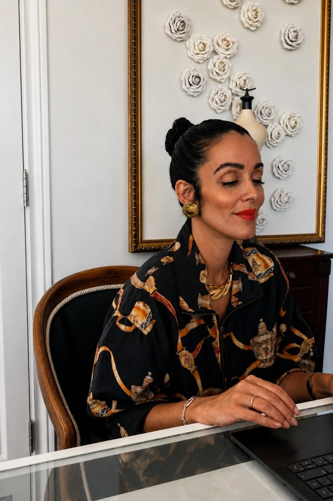 A woman with dark hair styled in a bun, wearing bright red lipstick, gold earrings, a patterned black shirt, and a bracelet, sitting at a desk with a laptop, in a room with a white wall, floral wall decor, and a small wooden table.