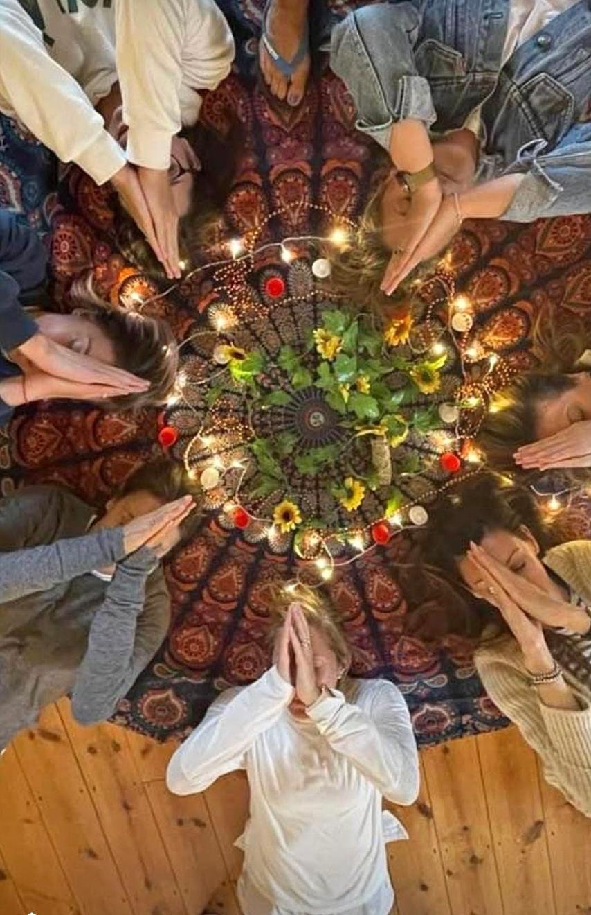 People gathered in a circle on a patterned carpet, praying with hands pressed together and heads bowed, with candles, sunflowers, and greenery in the center.