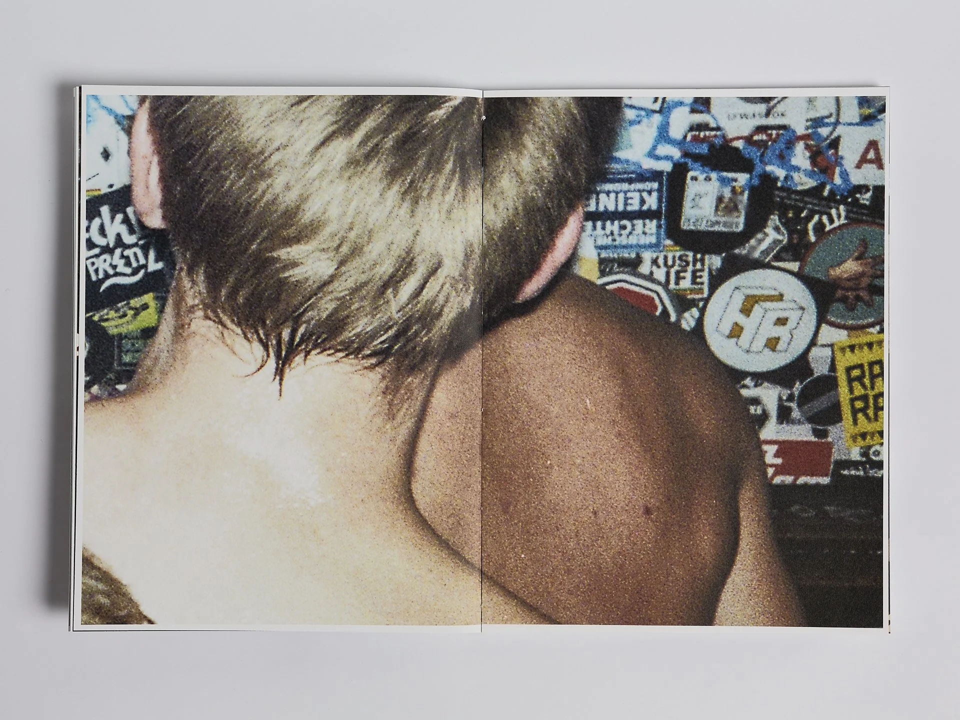 Ein offener Fotoband zeigt die Rückseite einer Person mit kurzen, blonden Haaren und nacktem Rücken, vor einer Wand mit zahlreichen bunten Aufklebern.