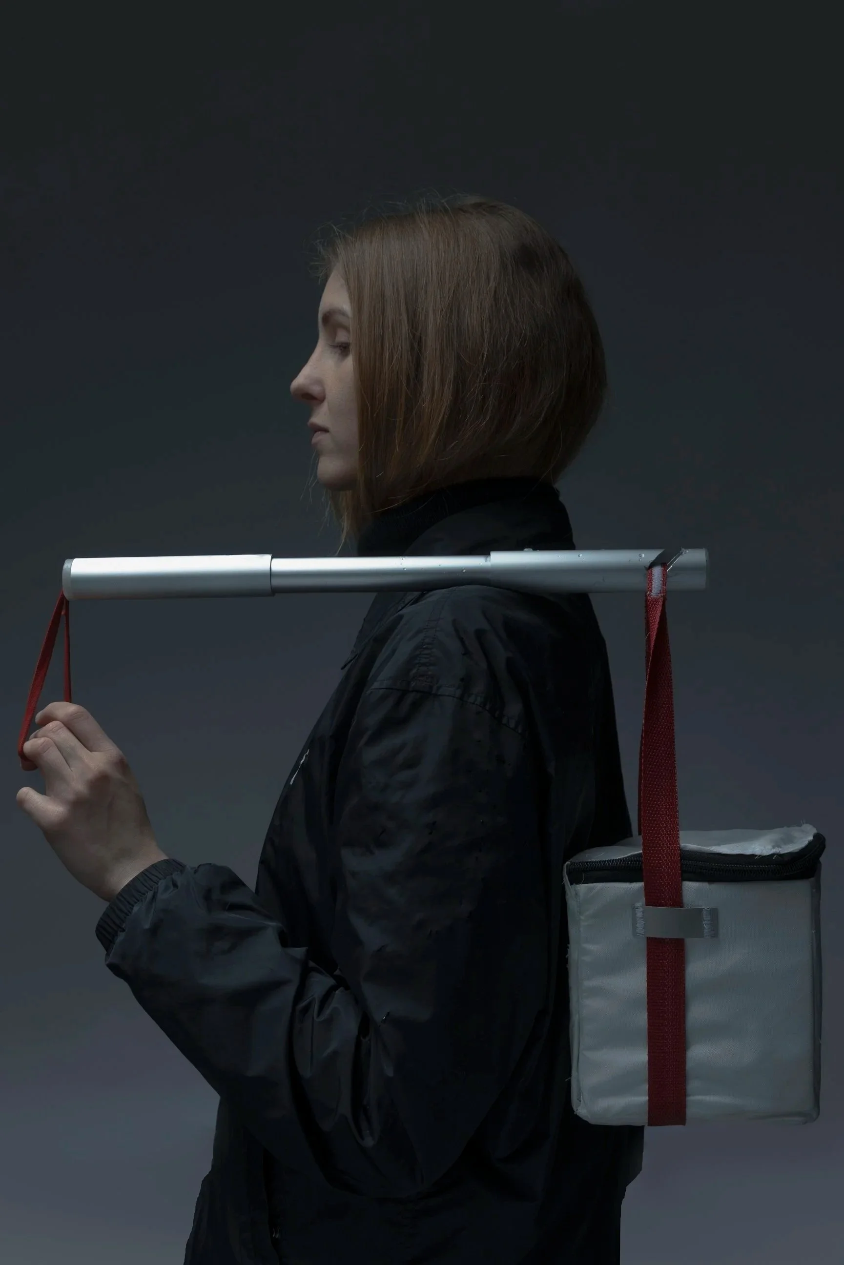 A woman with shoulder-length brown hair, dressed in a black jacket, is holding a bindle with red straps over her shoulders, and a metallic stick with red straps is resting on her shoulders. The background is dark and plain.