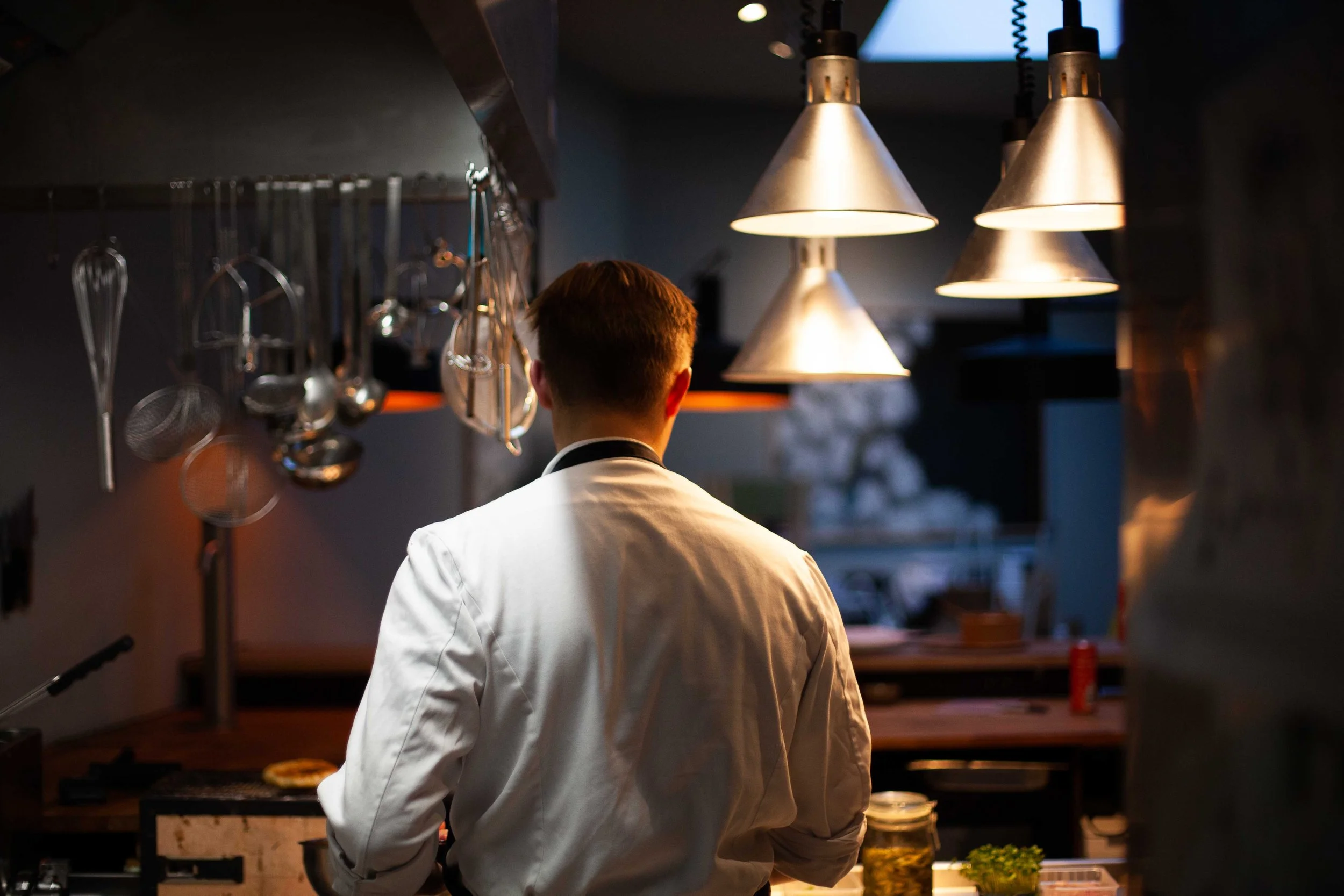 Küchenchef in einer Restaurantküche, der mit seinem Rücken zur Kamera steht, während er die Küche inspiziert, mit Teelichtern und Küchenutensilien im Hintergrund.