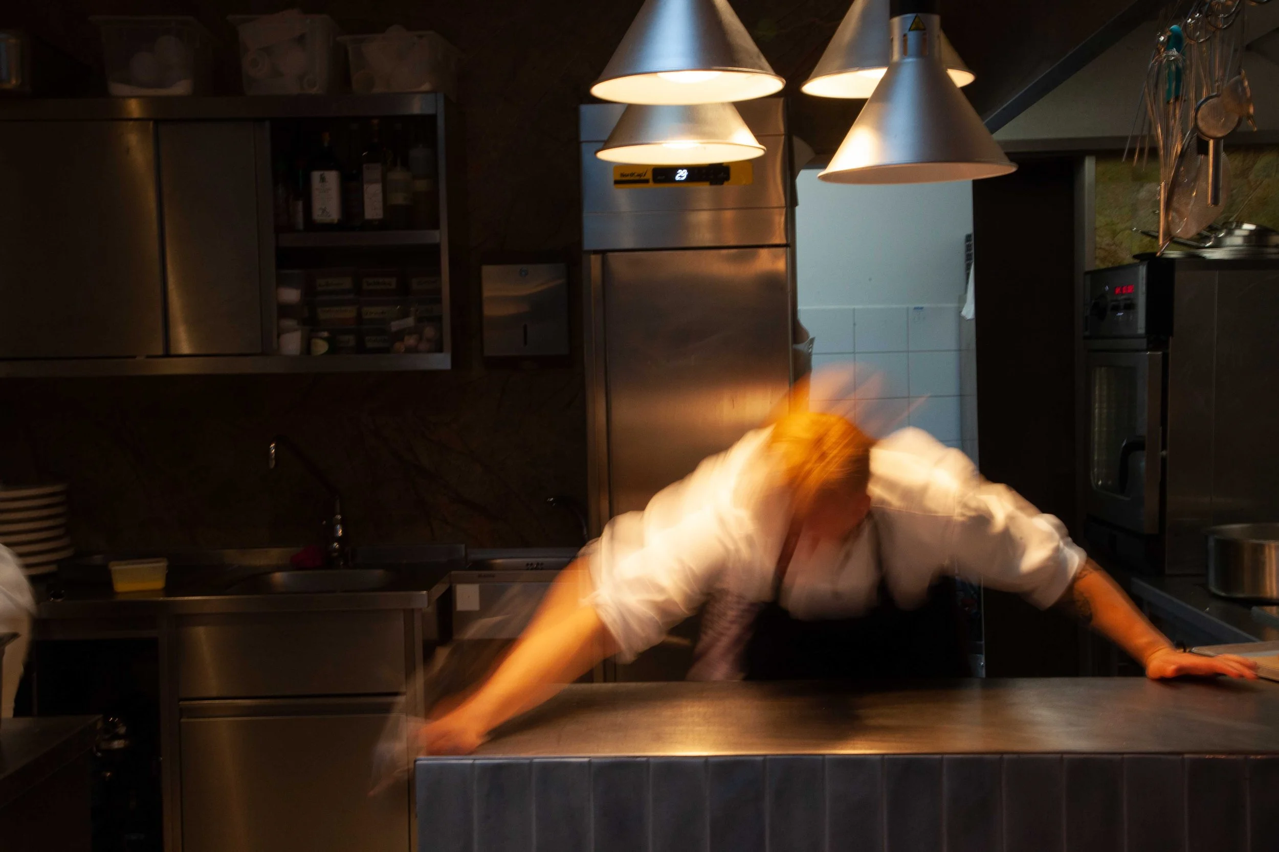 Ein Küchenbild mit dunklem Hintergrund, Metallregalen und einer Deckenlampe. Eine Person ist in Bewegung, wasunschwer erkennbar ist.
