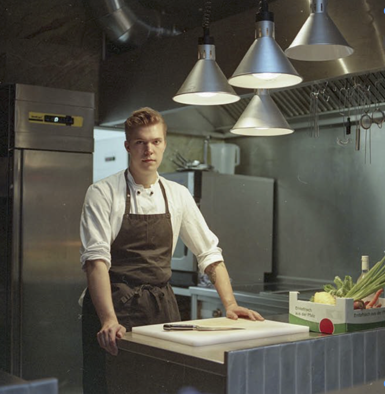 A young male chef stands behind a kitchen counter with a cutting board, in a commercial kitchen with hanging lights and stainless steel appliances, including a refrigerator and industrial vent.