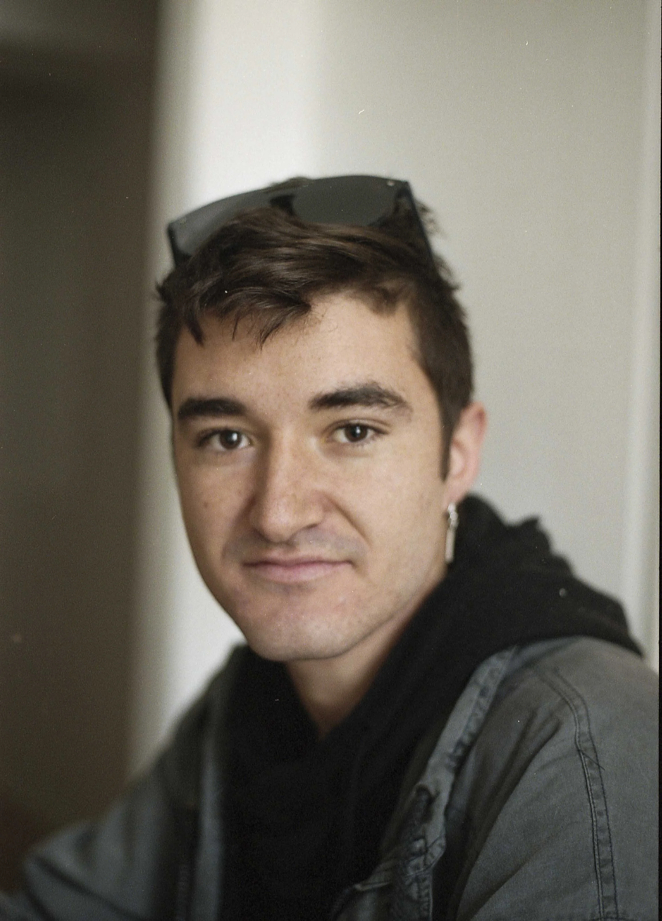 Ein junger Mann mit kurzen braunen Haaren trägt schwarze Sonnenbrillen auf dem Kopf, schwarze Jacke mit Kapuze und graues T-Shirt, blickt in die Kamera, im Hintergrund eine weiße Wand.