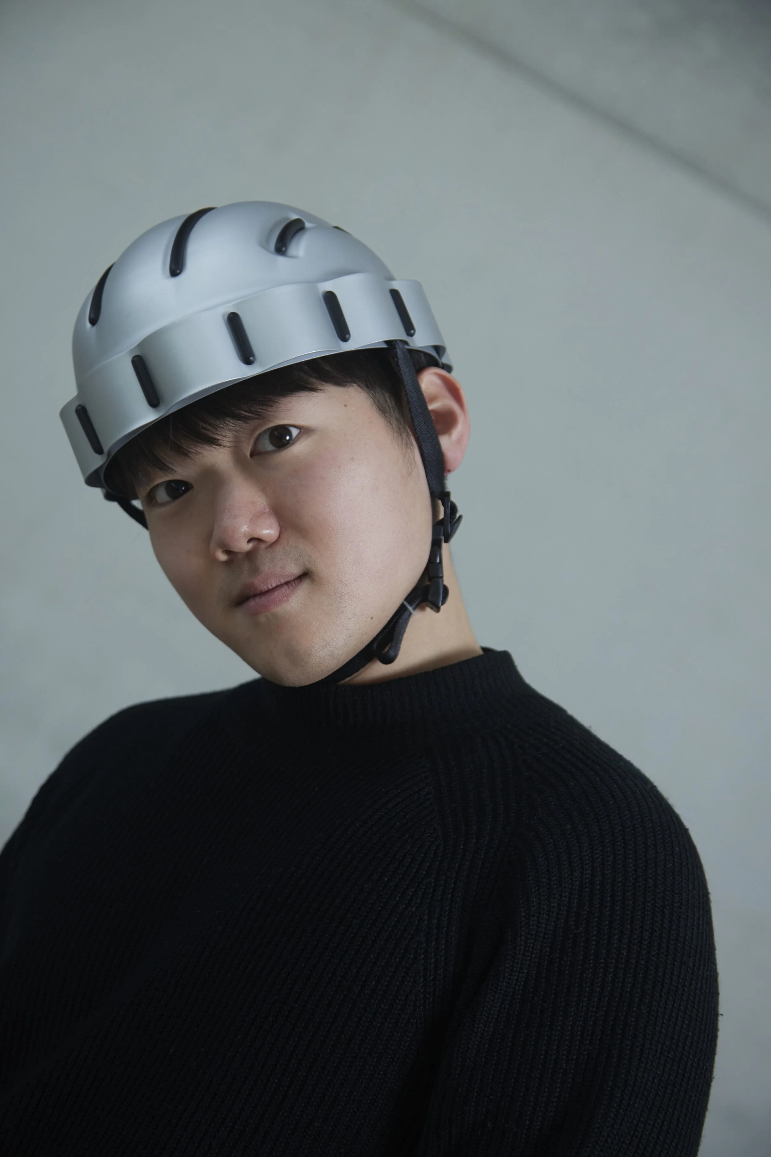 Nahaufnahme eines jungen Mannes mit dunklen Haaren, der eine weiße Kletterhelm trägt, vor einem neutralen grauen Hintergrund.