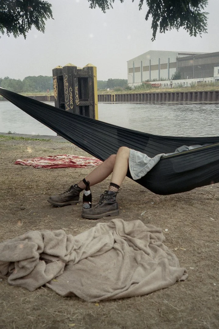 Person lying in a hammock by a body of water, with legs and boots visible, a blanket on the ground, and a large industrial building in the background.