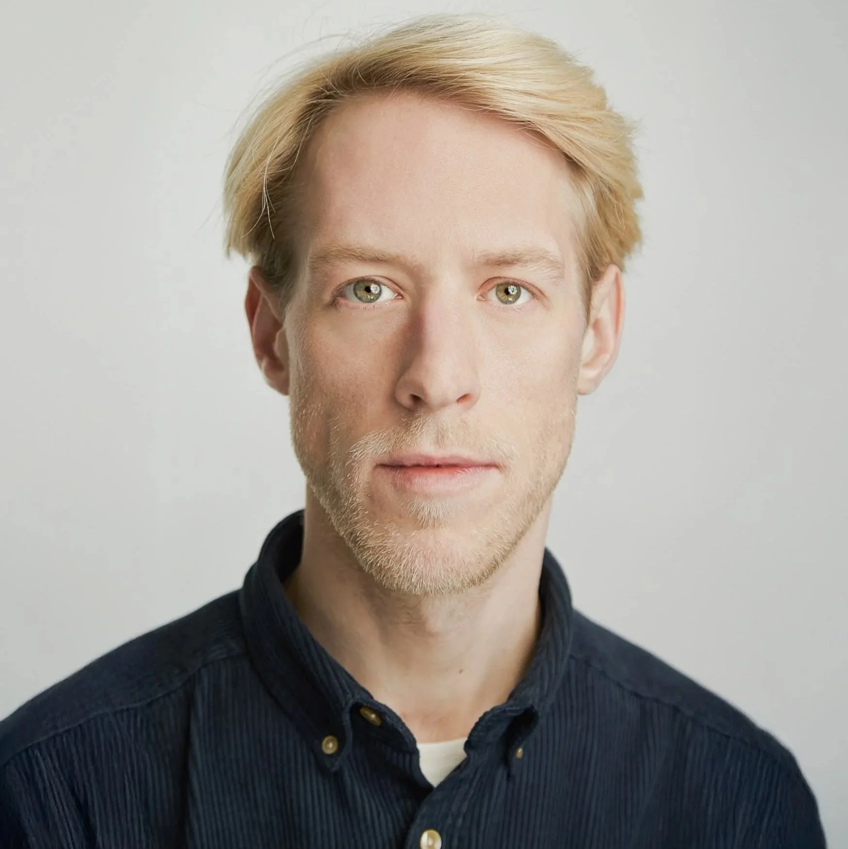 Porträt eines jungen Mannes mit blondem, seitlich gekämmtem Haar, blauen Augen, glattem Gesicht und hellen Haut, getragen mit einer schwarzen, hochgekrempelten Jacke vor einem weißen Hintergrund.
