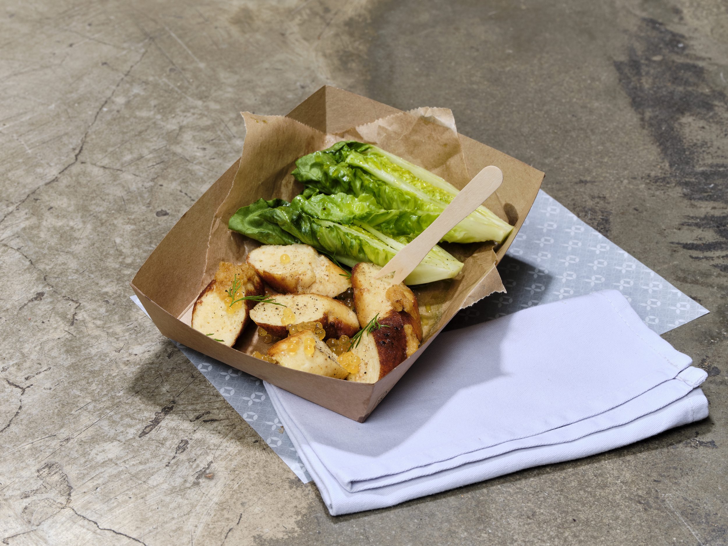 Frische grüne Salatblätter in einer braunen Pappschale, daneben gebratene Hähnchenstücke mit einer Sauce und kleinen kugelartigen Beilagen, auf einem weißen Unterteller mit Stoffserviette auf einer Steinoberfläche.