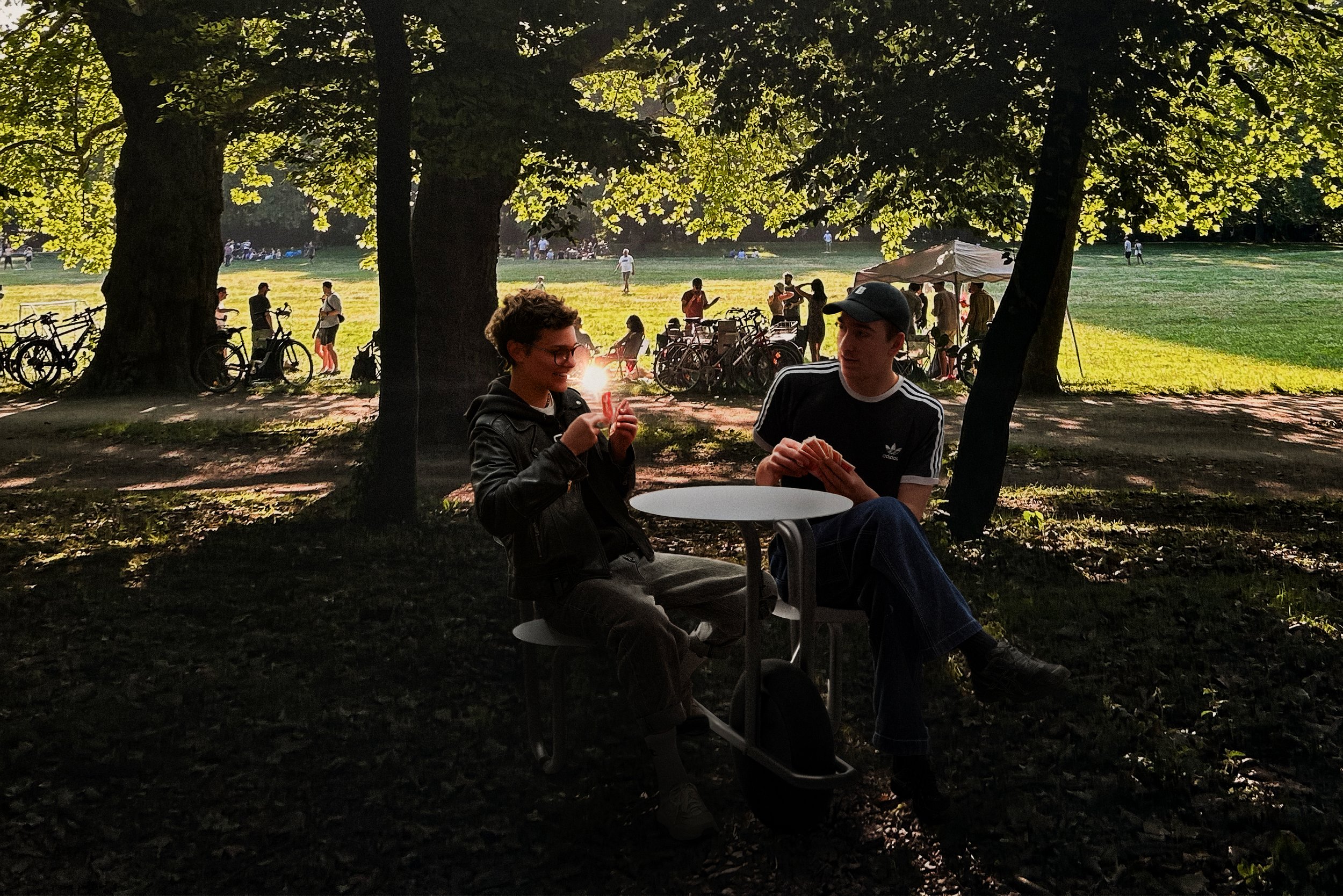 Zwei junge Menschen sitzen im Schatten unter Bäumen in einem Park, im Hintergrund sind weitere Menschen, Fahrräder und ein Zelt. Es ist sonnig und die Atmosphäre wirkt entspannt.