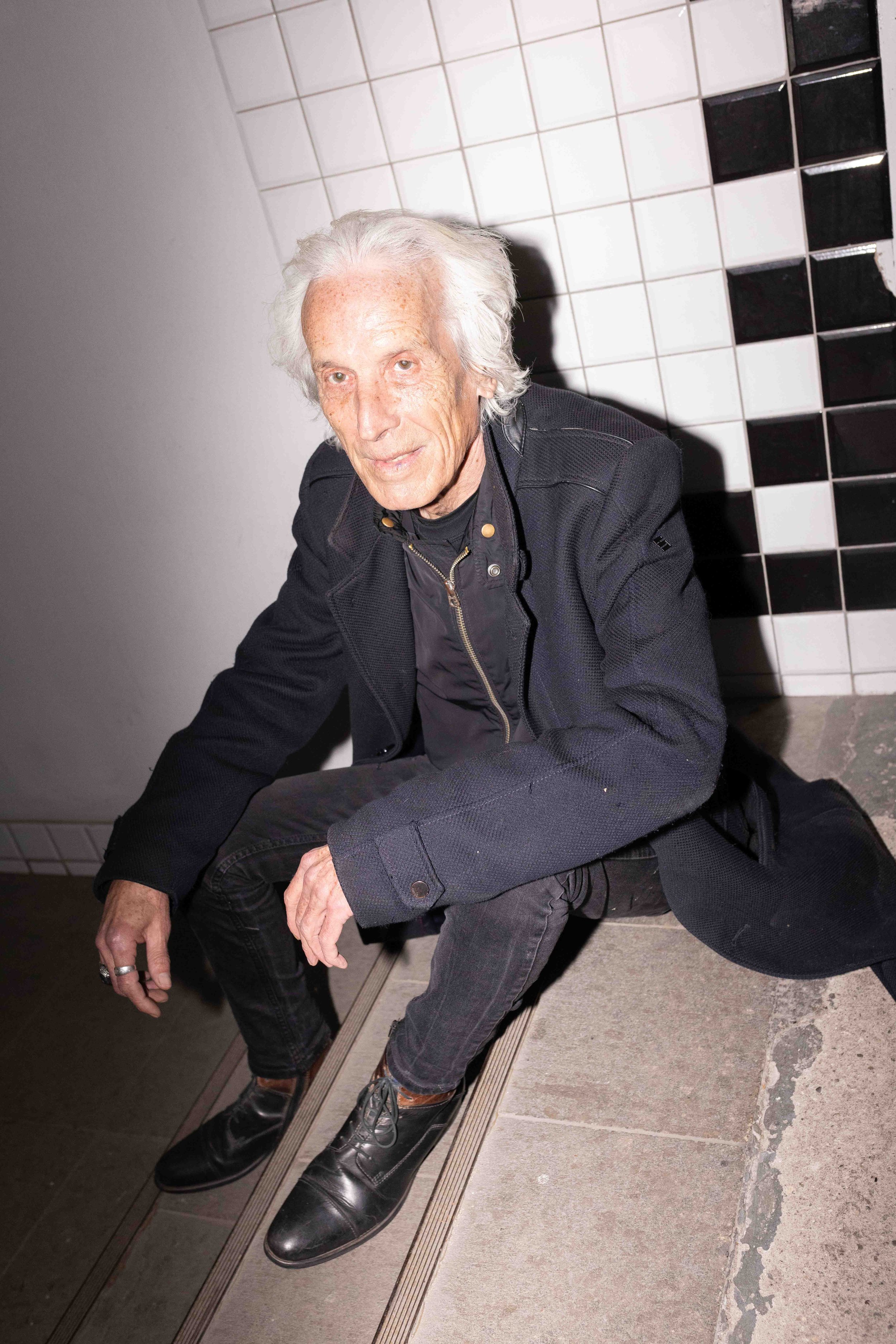 Ein älterer Mann mit weißen Haaren sitzt auf dem Boden vor einer Wand mit schwarz-weißen Fliesen, trägt eine schwarze Jacke und schwarze Schuhe.
