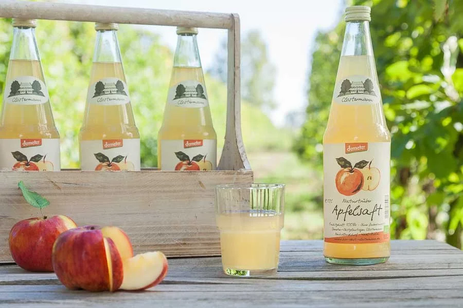 Eine Flasche Apfelsaft, ein Glas mit Apfelsaft, drei Äpfel, zwei davon halbiert, auf einem Holztisch, im Hintergrund eine Holzkiste mit vier weiteren Flaschen Apfelsaft vor grünen Bäumen.