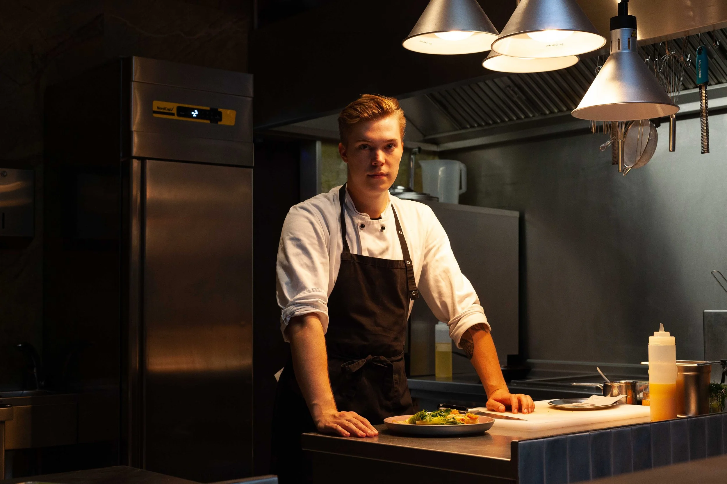Ein junger männlicher Koch in einem Restaurantküche, der vor einem Teller mit frischem Salat steht. Über ihm hängen metallene Lampen und es sind verschiedene Küchengeräte und Zutaten sichtbar.