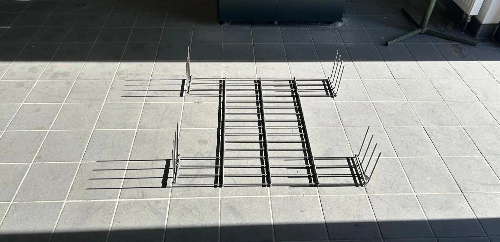 Drahtgestell auf Bodenfliesen, Schatten im Sonnenlicht.