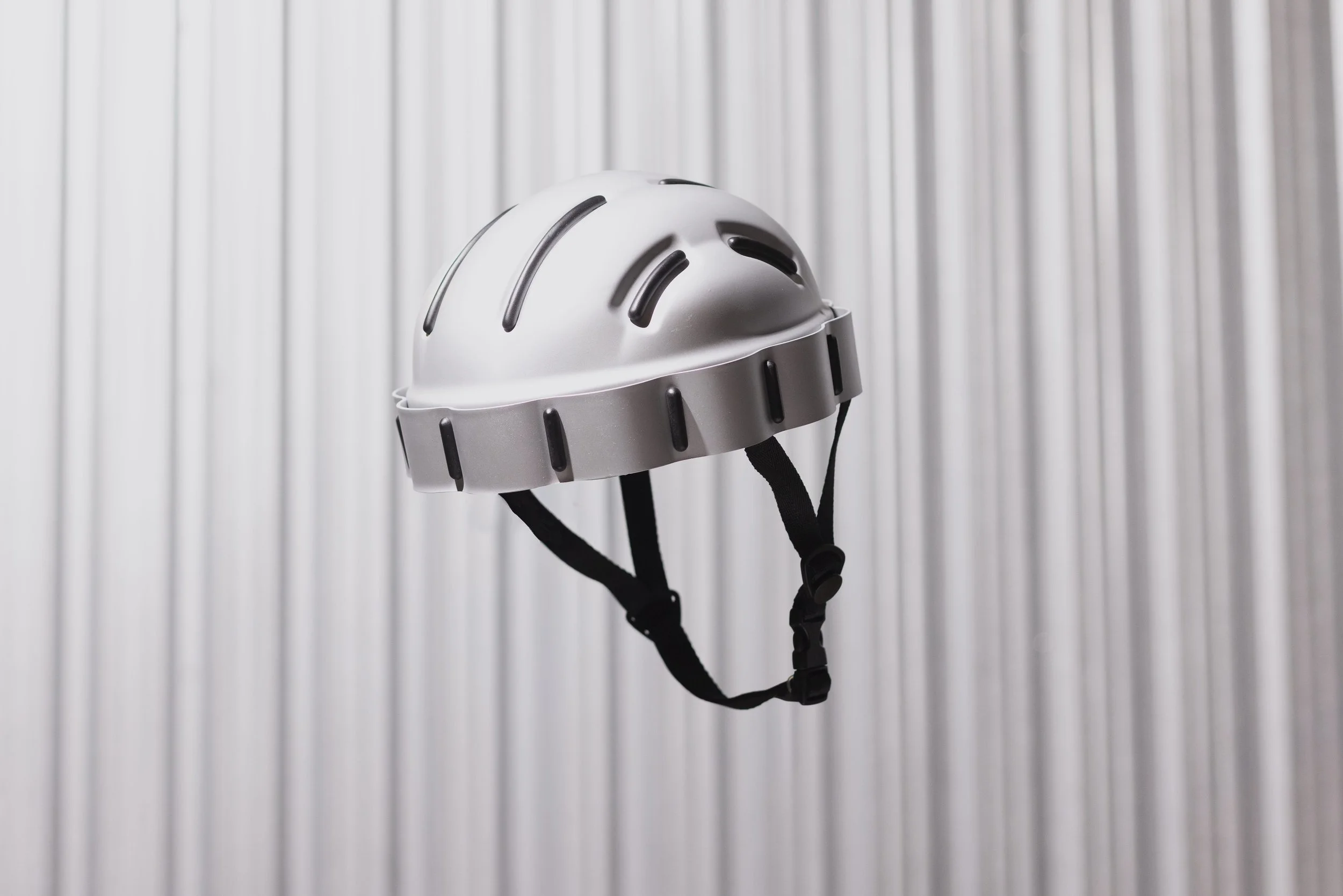 Ein weißer Fahrradhelm mit schwarzen Belüftungsöffnungen, der an einem schwarzen Kinnriemen hängt, vor einem vertikalen, silbernen Metallschrank.