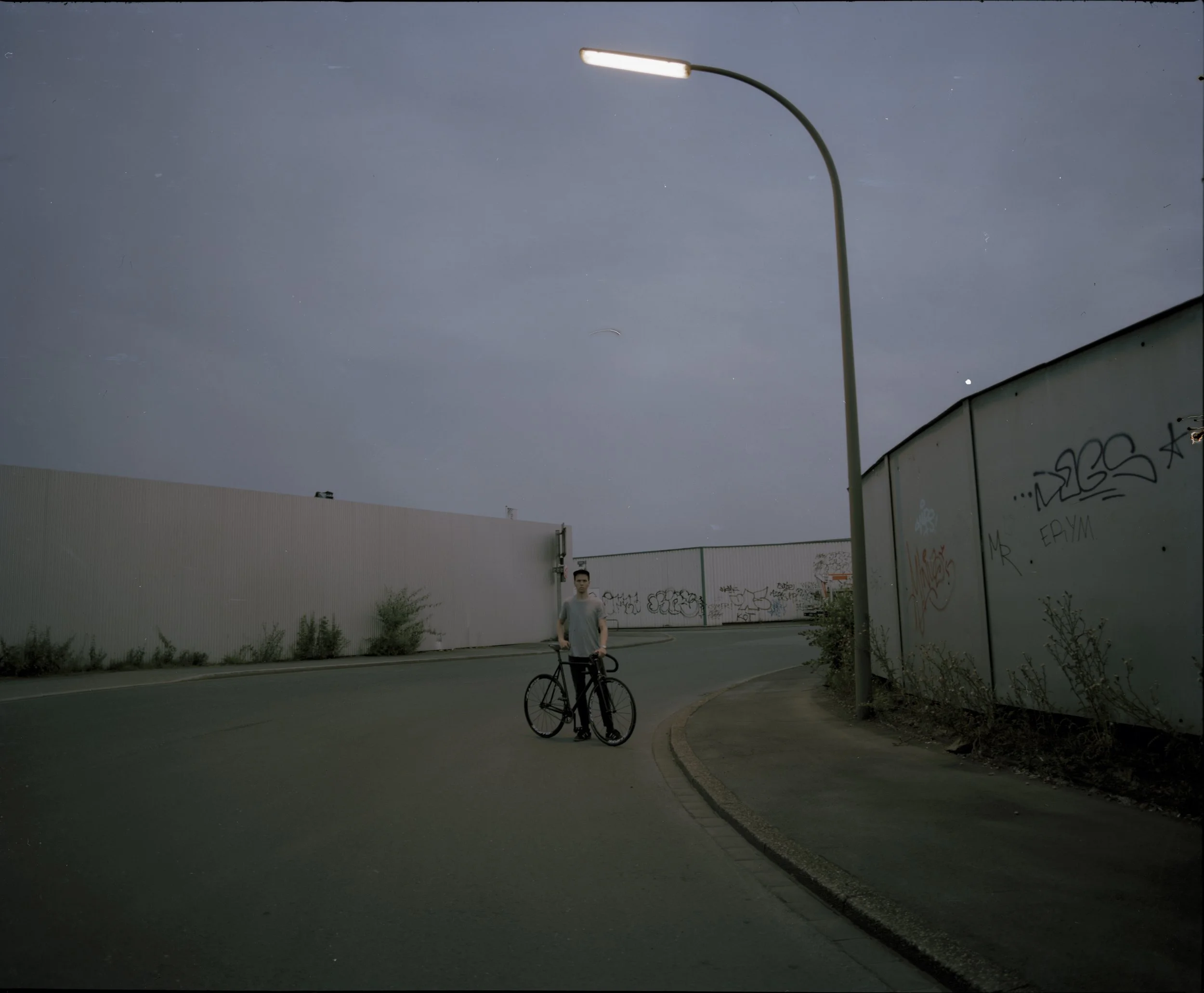Ein junger Mann mit Fahrrad an einer städtischen Straße bei Dämmerung, Graffiti an der Wand, Himmel ist grau und bewölkt.