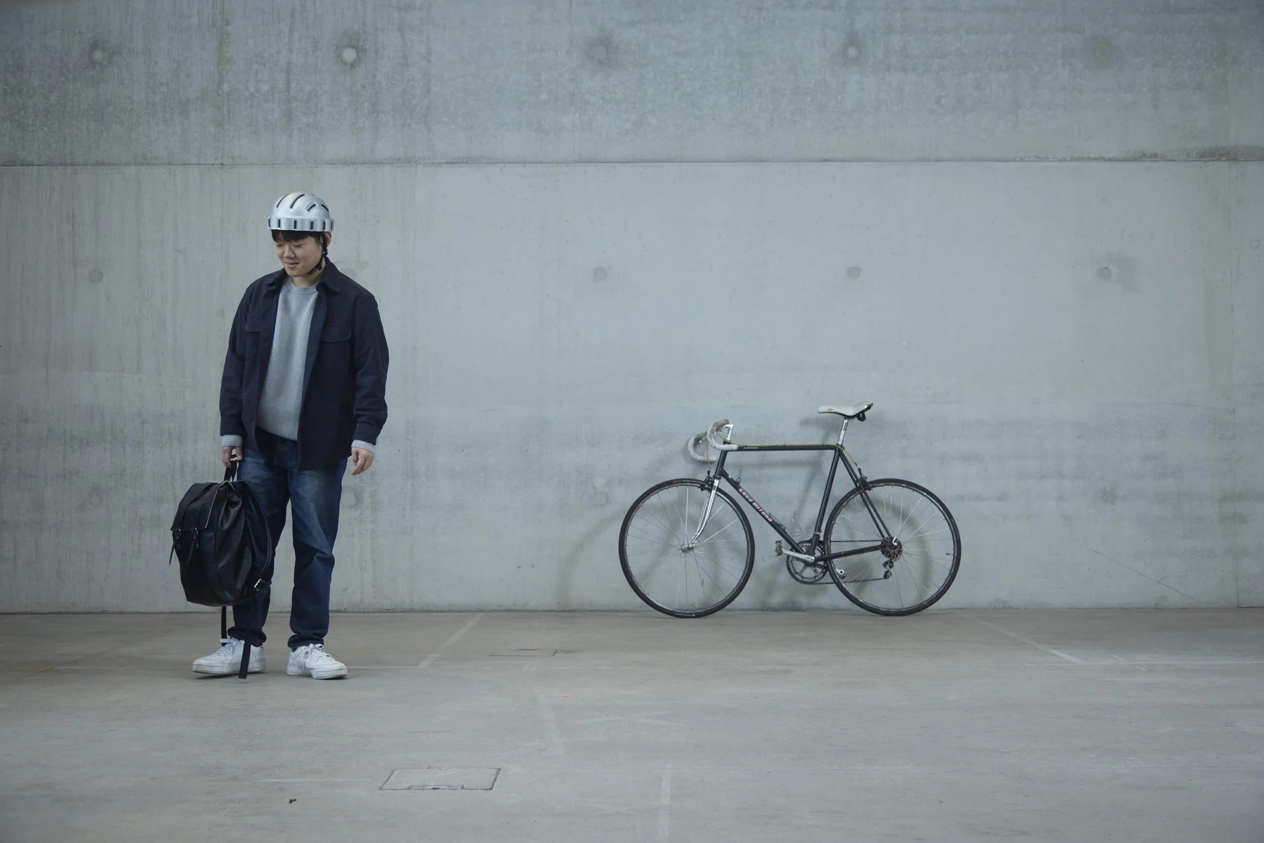 Ein junger Mann steht mit einem Rollstuhl, eine Radtasche und einer Fahrrad, vor einer grauen Betonwand.