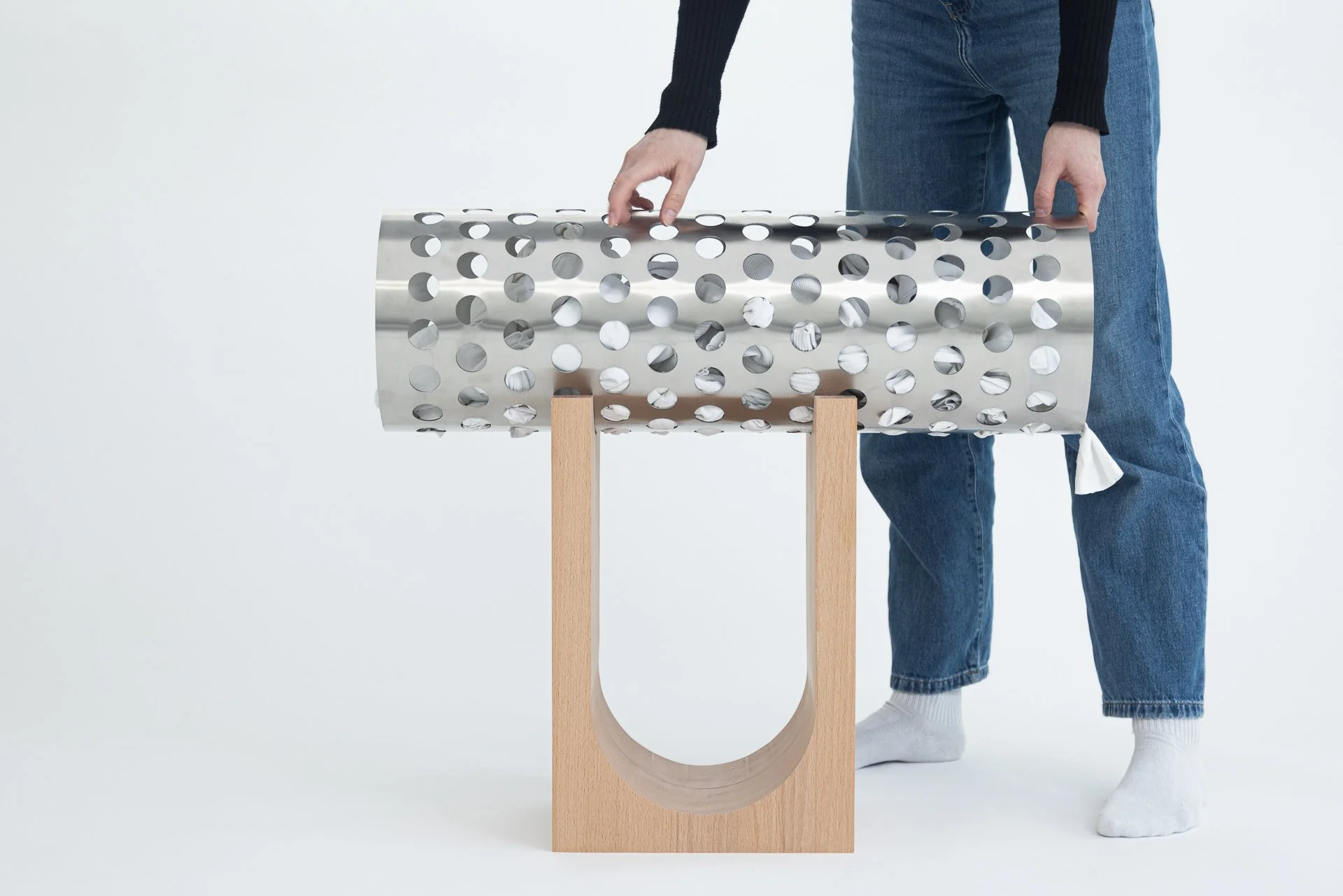 Person in Jeans und Pullover, die eine metallene, perforierte Röhre auf einem Holzgestell hält, Werkzeuge im Hintergrund, weißer Hintergrund.