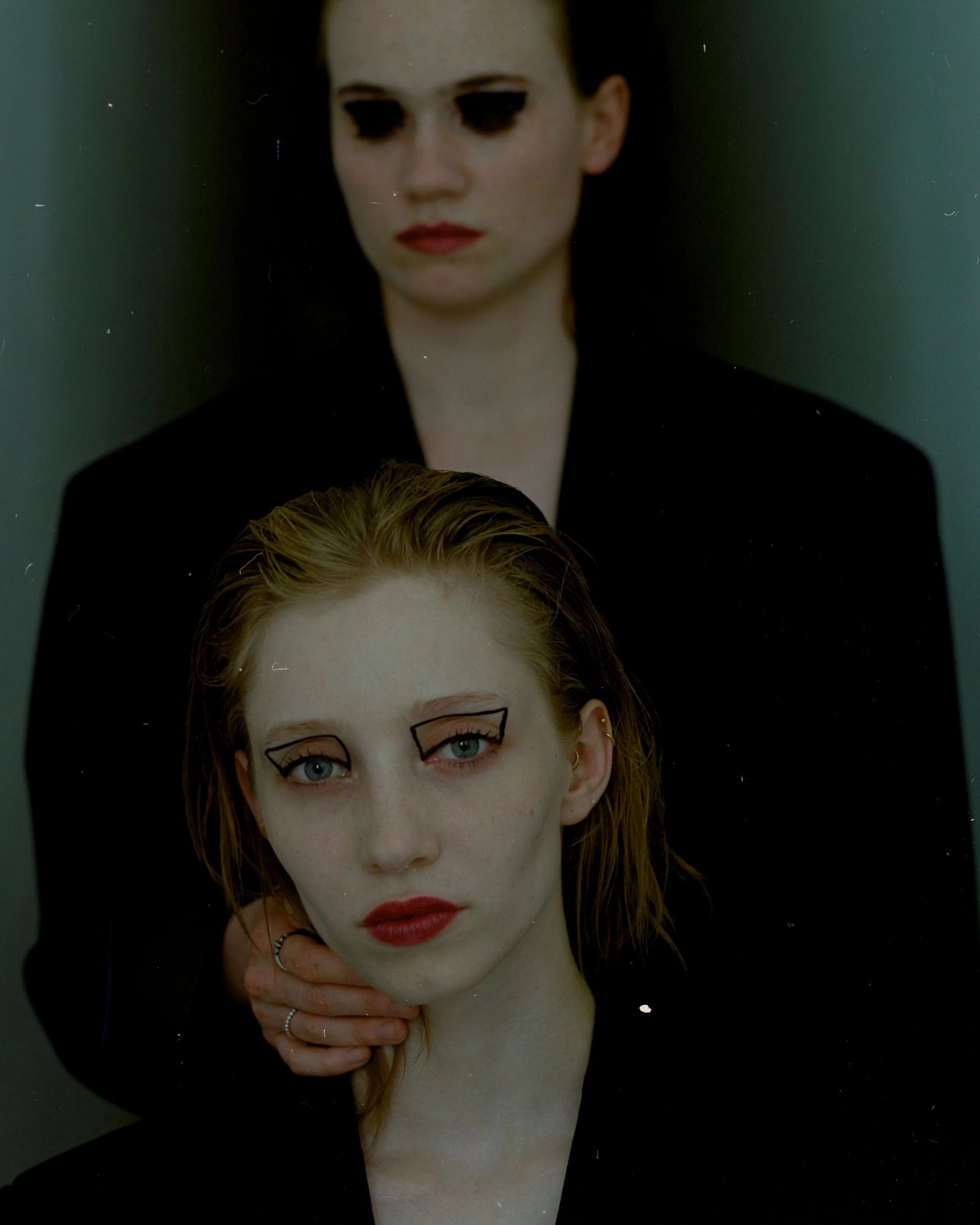 Doppelporträt eines jungen Mädchens mit roten Haaren, glattem Haar, blauen Augen, Make-up mit grüner Kontur auf den Augen, rotem Lippenstift, tragen eine schwarze Jacke, auf dem Bild befindet sich eine Hand, die das Mädchen hält.