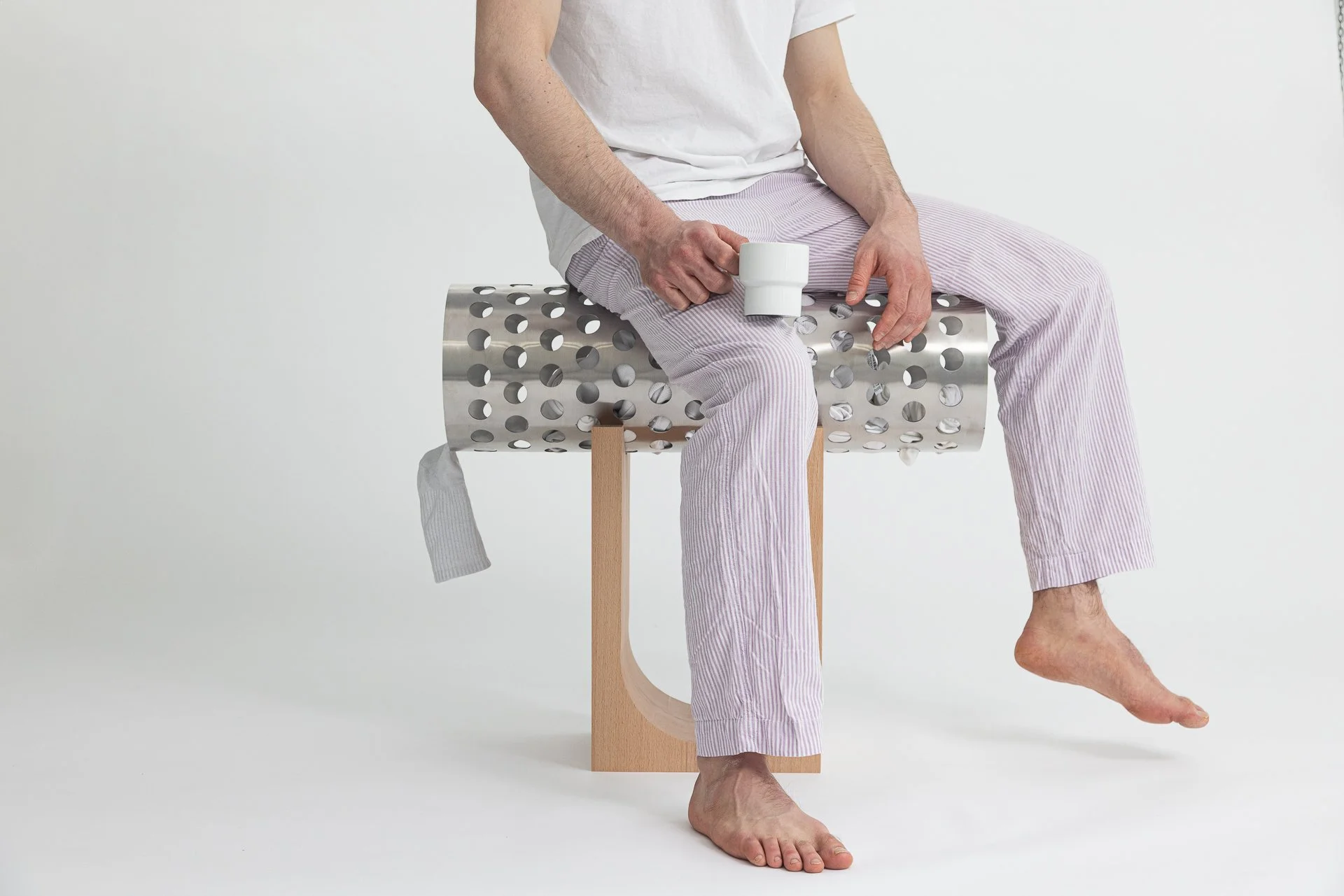 Ein Mann sitzt auf einem weißen, perforierten Metallhocker, trägt weiße und lila gestreifte Schlafhose sowie ein weißes T-Shirt, hält einen weißen Becher in der Hand und hat die Füße auf dem Boden während er das rechte Bein hochgestreckt hat.