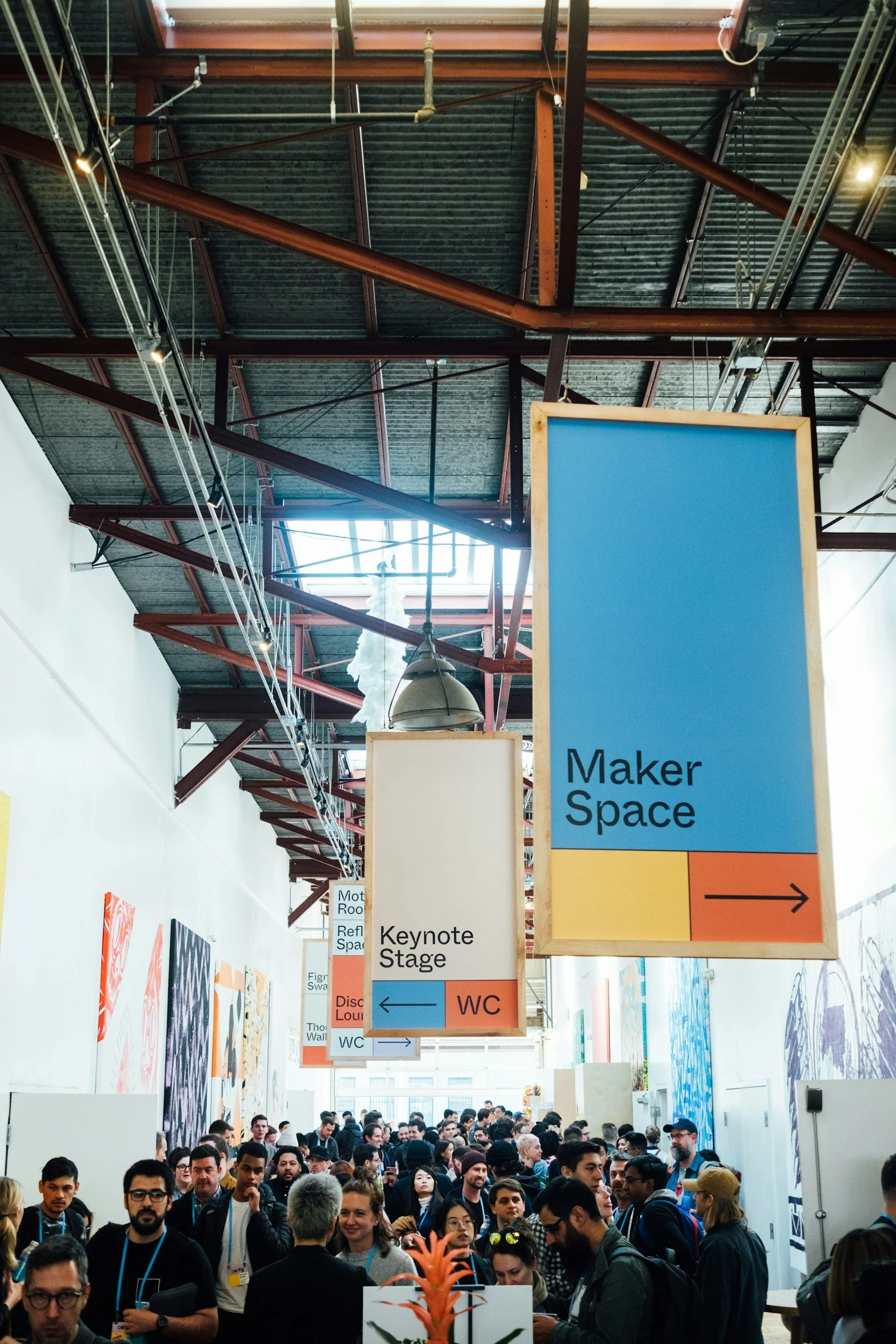Crowd of people in a large indoor space with signs indicating directions to a Maker Space, keynote stage, and restrooms. The ceiling has exposed metal beams and industrial lighting.