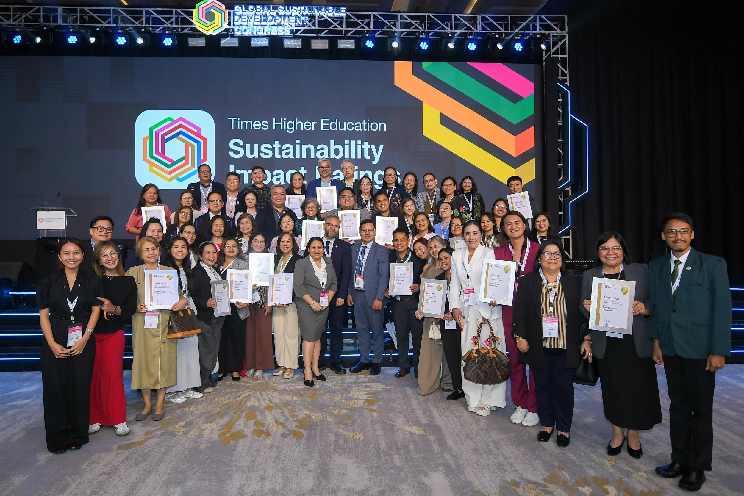 A large group of people, dressed professionally, gathered on a stage at the Global Sustainable Development Congress. Some are holding certificates and smiling. The stage features a big screen displaying the event’s logo, which includes colorful interconnected hexagons, along with the phrases 'Times Higher Education' and 'Sustainability Impact Ranking'.