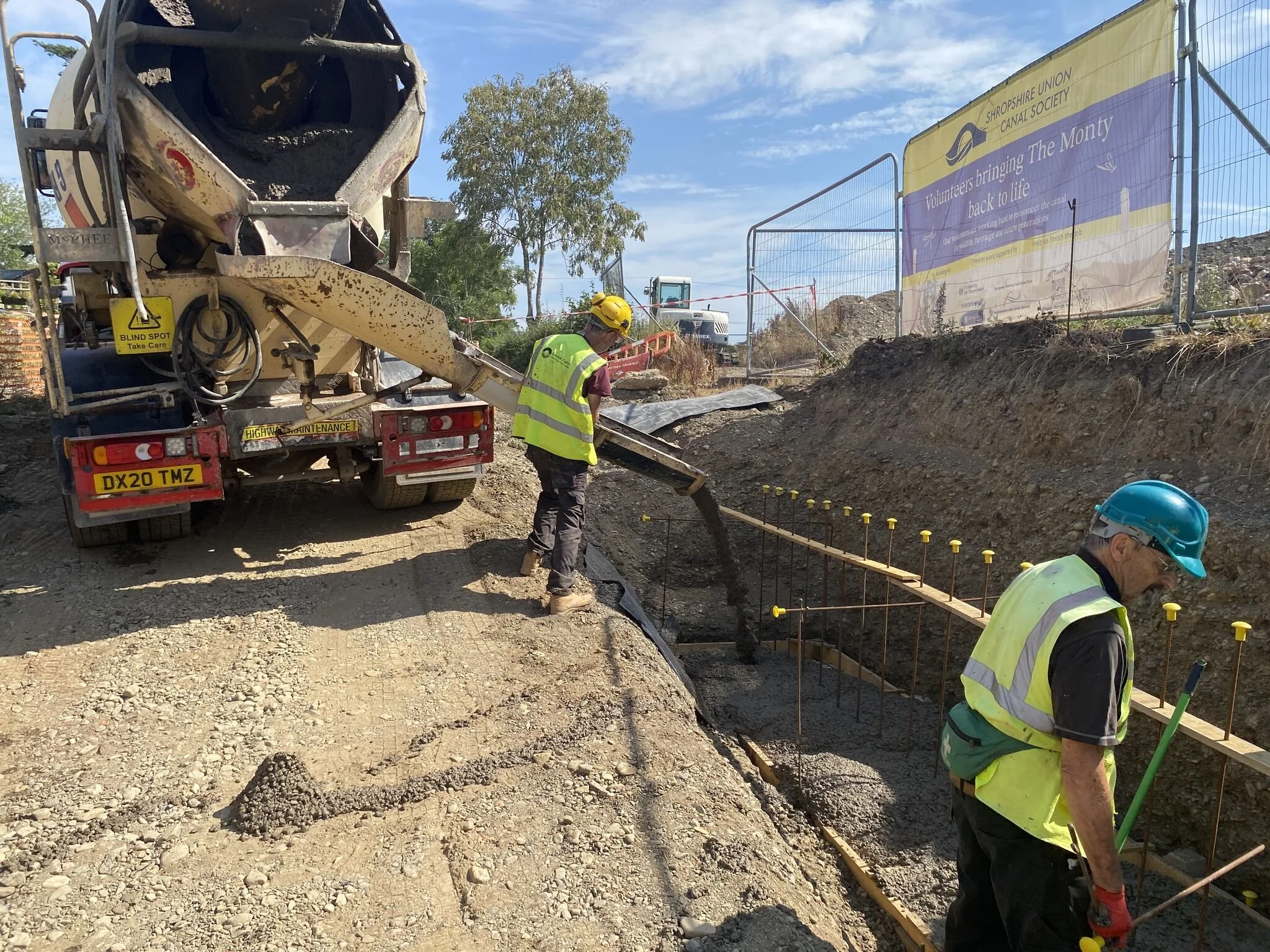 Construction workers in high-visibility vests and helmets working on a concrete foundation at a construction site with a cement mixer truck to the left and a sign for the Shropshire Union Canal Society to the right.