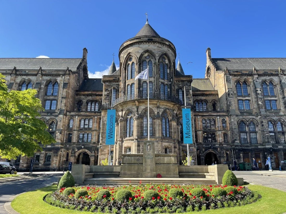 The front of a historic stone building labeled as a museum with banners saying 'Welcome' and 'Museum' on either side of a fountain with a flag. There is green grass, flowers, and trees in front of the building.