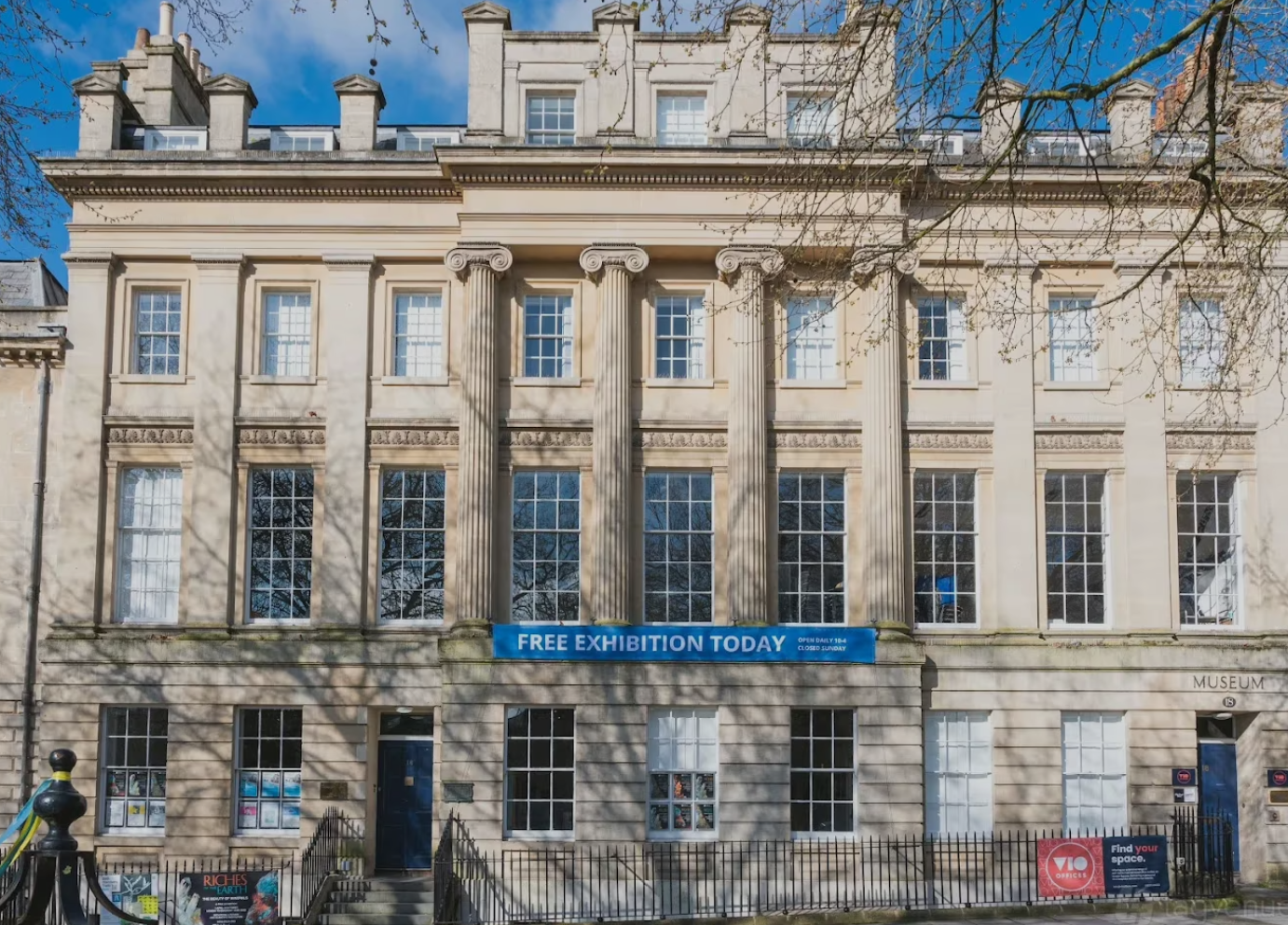 Facade of a historic building with large windows, decorative columns, and a blue banner advertising a free exhibition today. The building has a classical architectural style, with a clear sky and tree shadows visible.