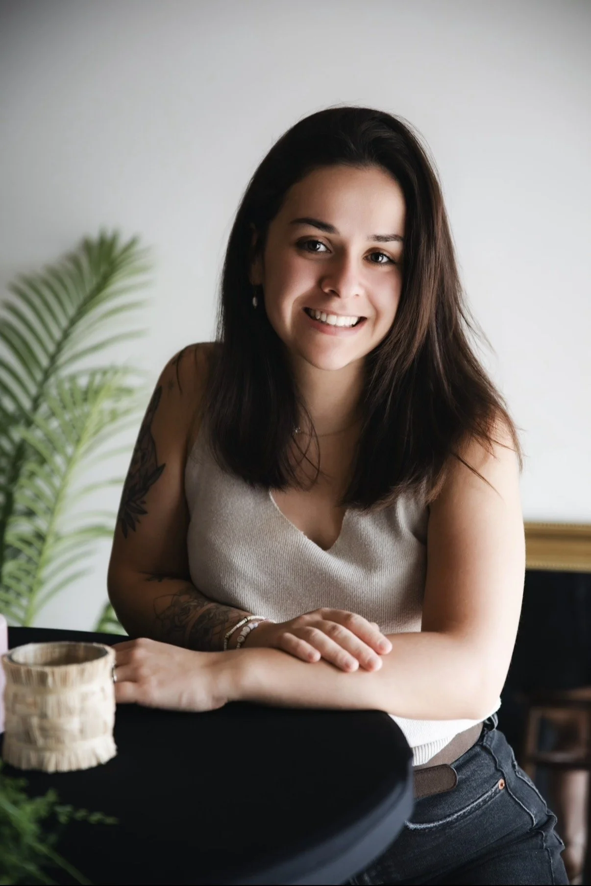 Jeune femme assise à une table, souriante, avec un tatouage sur le bras, portant un haut beige.