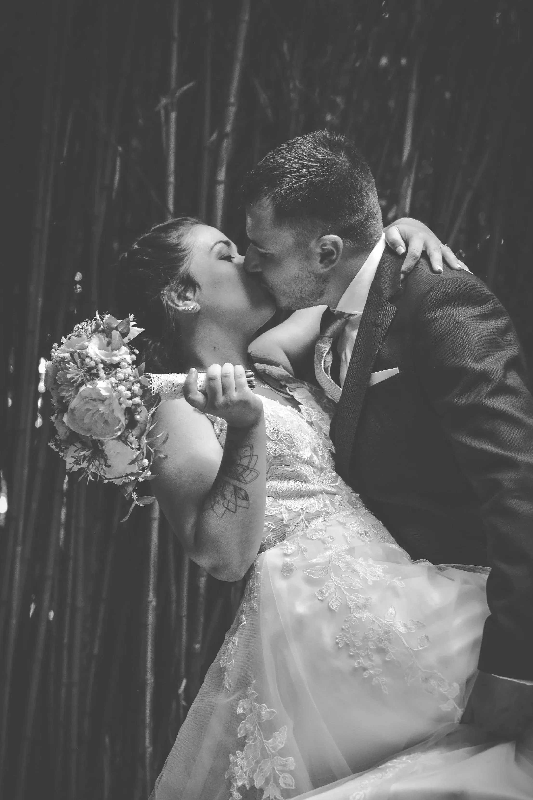 Un couple de mariage s'embrasse dans un cadre romantique, la femme porte une robe de mariée avec des broderies florales et tient un bouquet, l'homme porte un costume et une cravate.