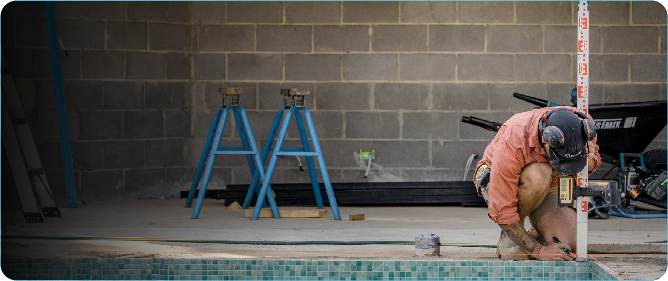 Construction worker wearing a black cap and orange shirt using a level and marker to measure near a swimming pool construction site.
