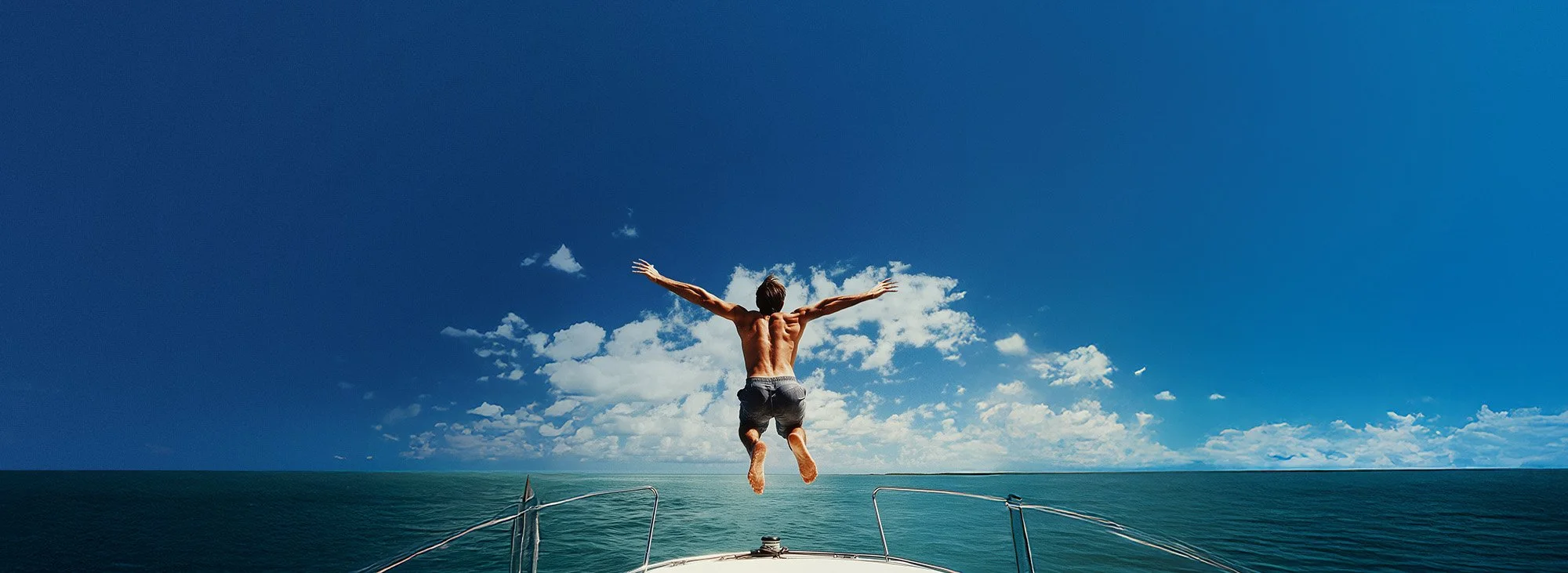 A man jumps into the water from a boat on a clear day with blue sky and scattered clouds.