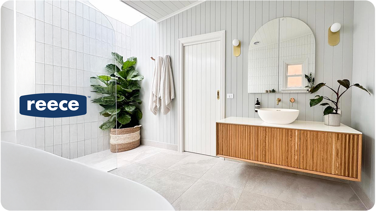 Modern bathroom with white tiled walls, a wooden vanity with a vessel sink, a large mirror, and a potted plant. There are two wall-mounted lights and a door, with a towel hanging on a hook. A large houseplant is in a woven basket near the corner.