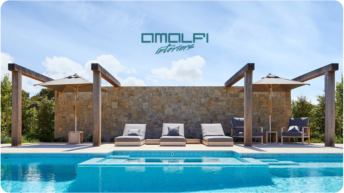 A luxurious outdoor pool area with lounge chairs, umbrellas, a stone wall, and a blue sky with clouds, featuring the text 'AMALFI interiors' at the top.