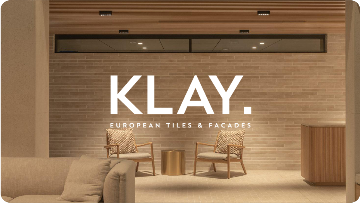 Interior of a modern lobby with two armchairs, a small gold side table, a beige sofa with pillows, and a brick wall with large illuminated sign reading 'KLAY. European Tiles & Facades.' ceiling lights and a large window or reflection above.